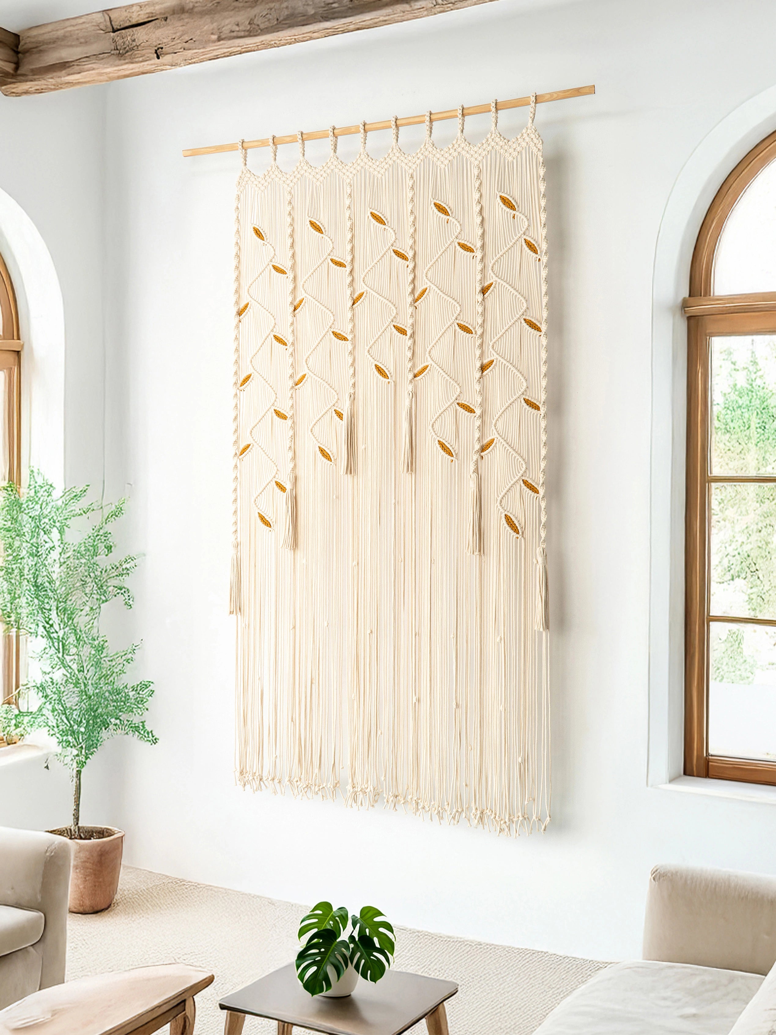 A large cream macrame wall hanging with geometric woven patterns and fringe, suspended from a wooden rod in a bright living room with arched windows and plants.