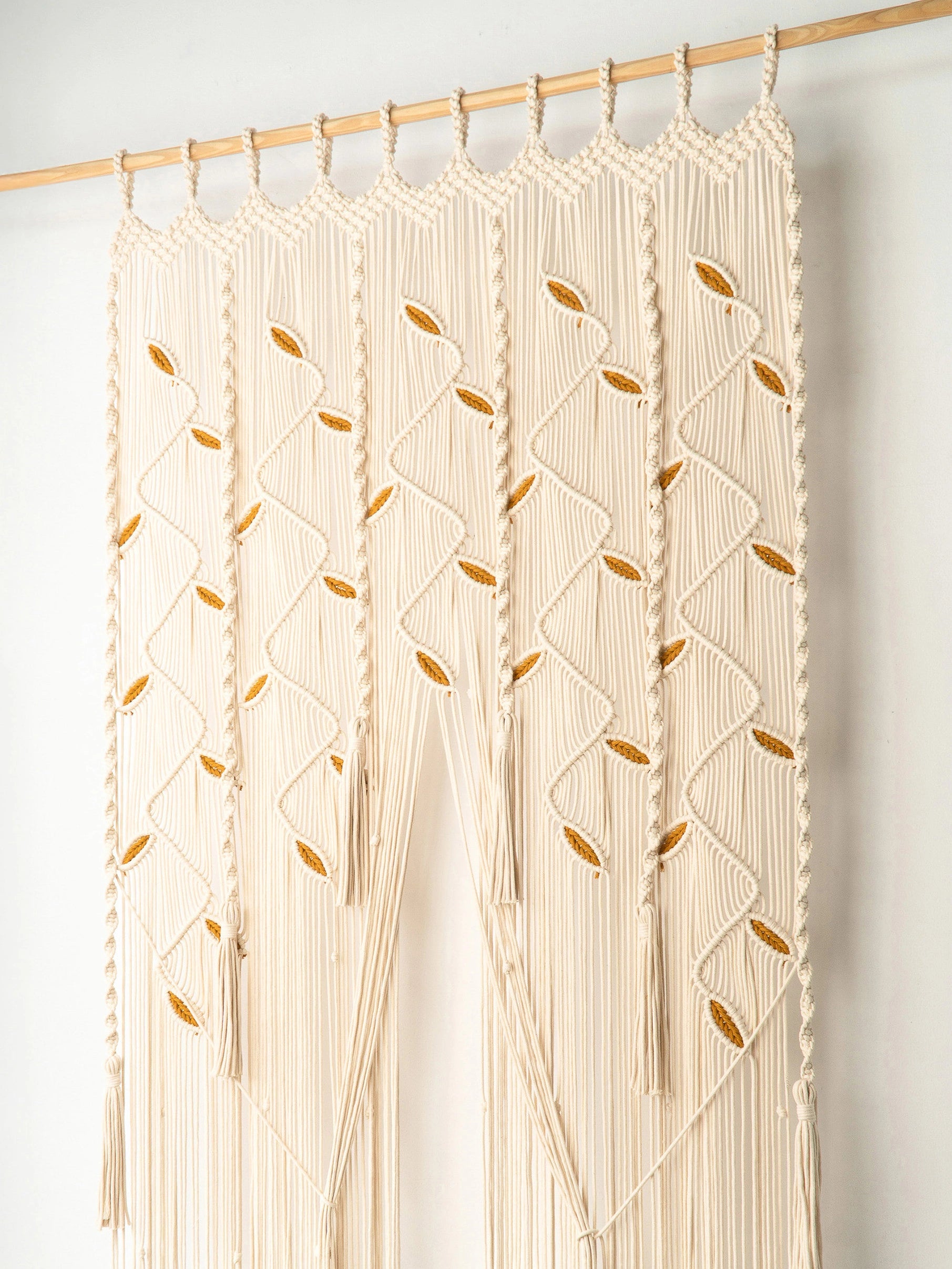 Cream macrame wall hanging with leaf patterns, suspended from a wooden rod against a plain white wall.