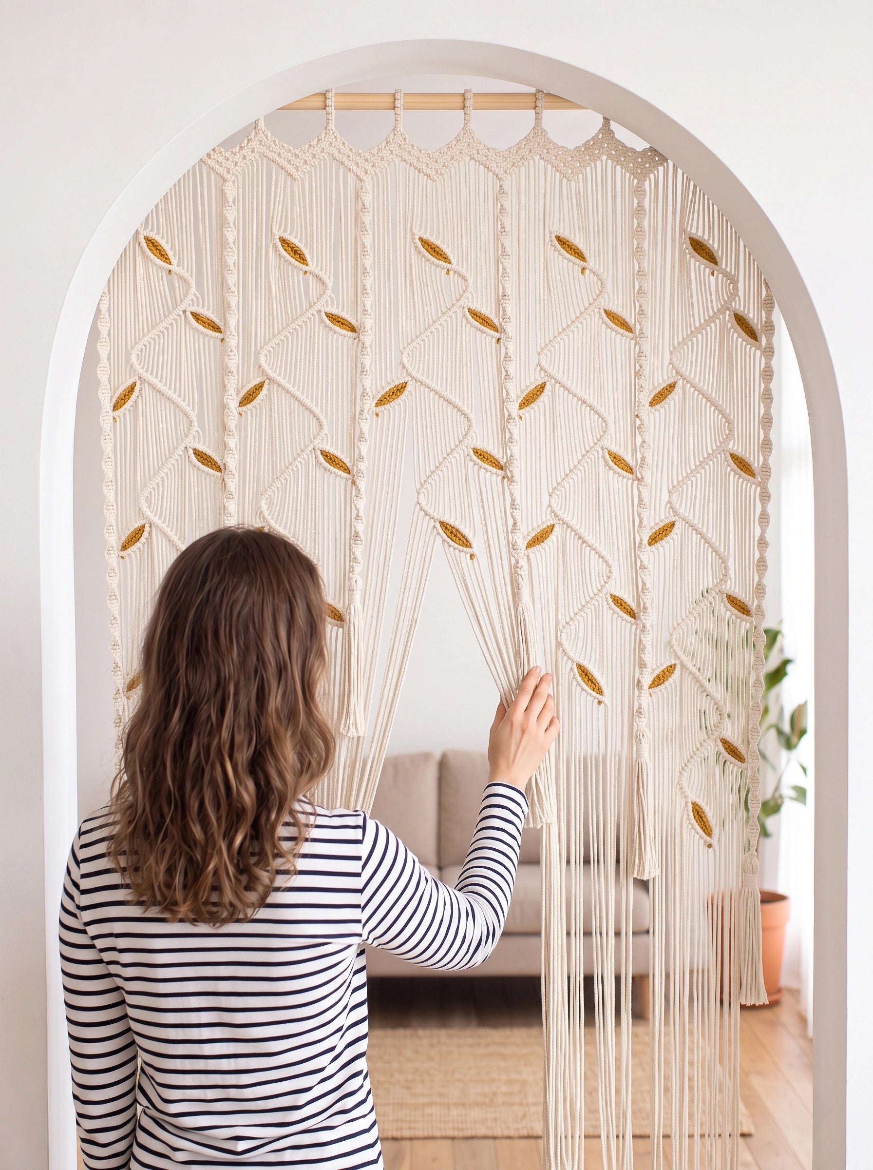 A woman in a black and white striped shirt stands in front of a large cream macrame curtain with gold-toned woven accents, holding one side of the hanging textile in a bright room with an arched doorway.