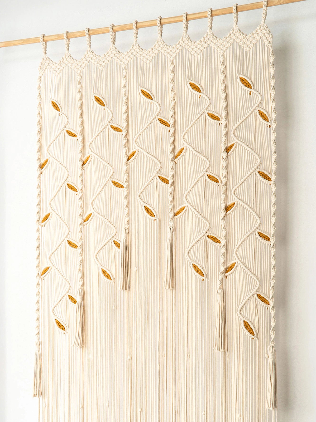 Cream macrame wall hanging with braided leaf patterns and long fringe, suspended from a wooden rod against a plain white wall.