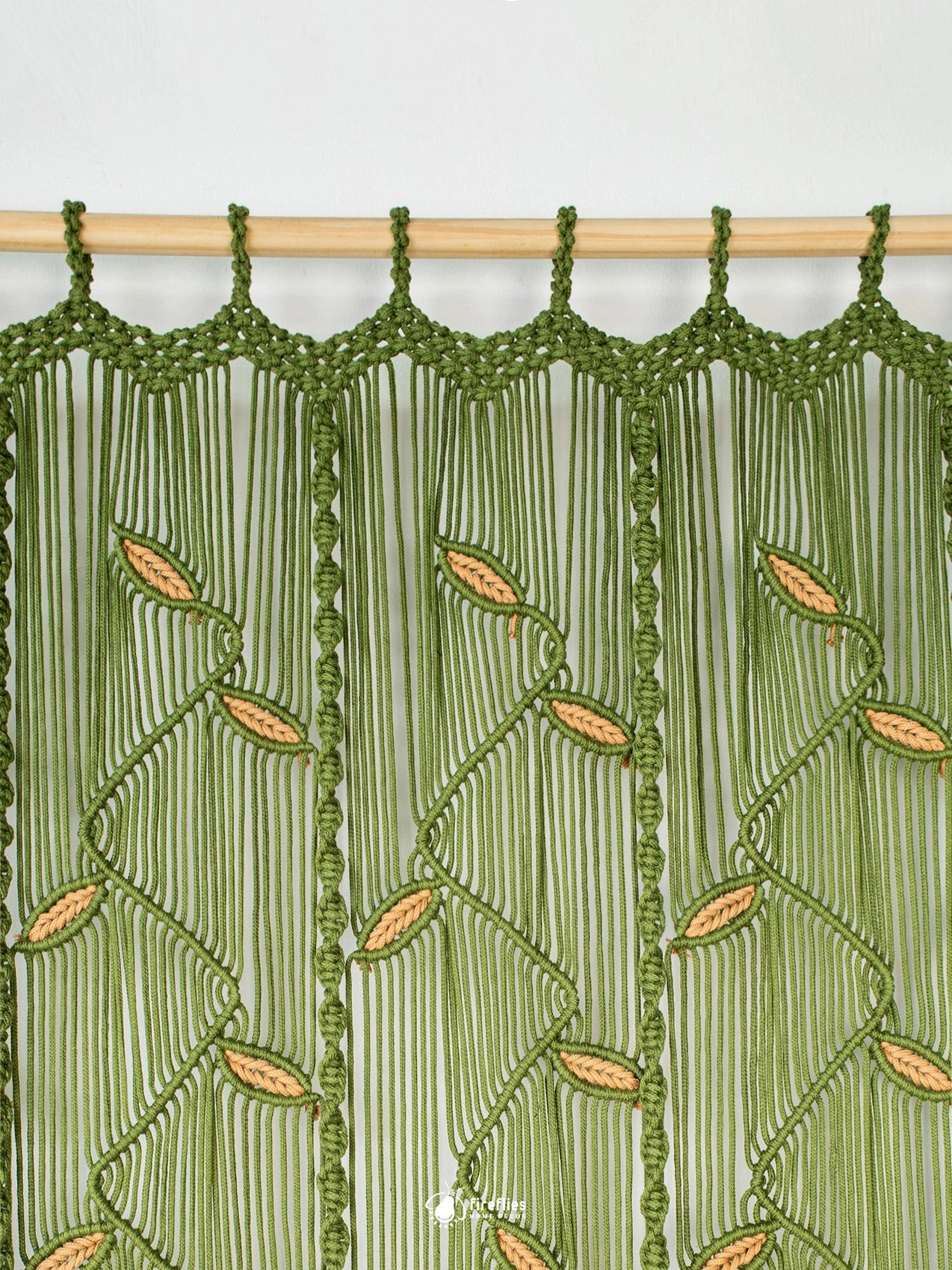 A close-up of a green macrame curtain panel hanging from a light wood rod. The woven design features vertical hanging cords with a repeating leaf pattern in a warm tan/gold color.