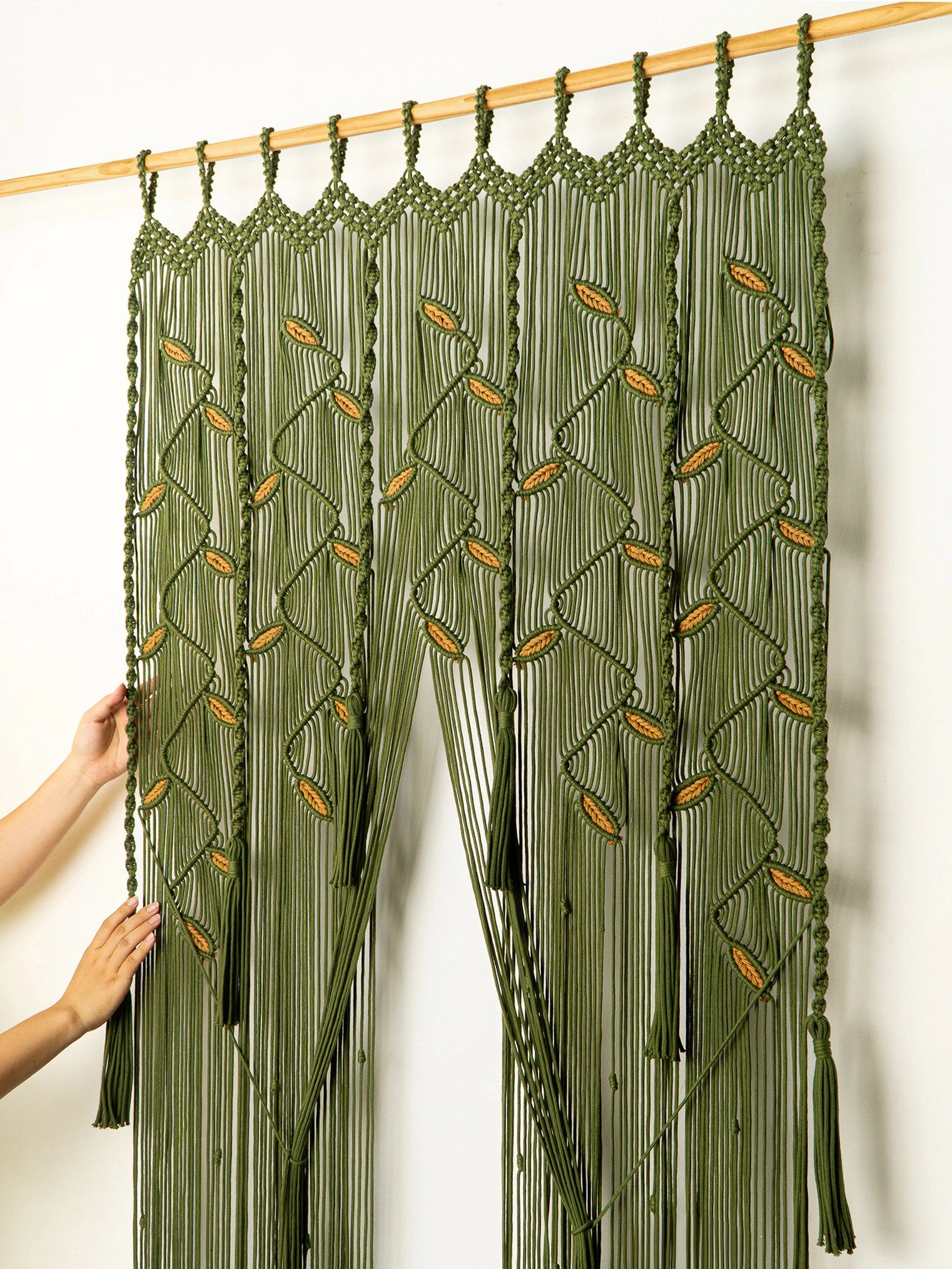 An olive green macrame curtain panel hanging from a wooden rod against a white wall, with a person's hands adjusting the left side of the panel.