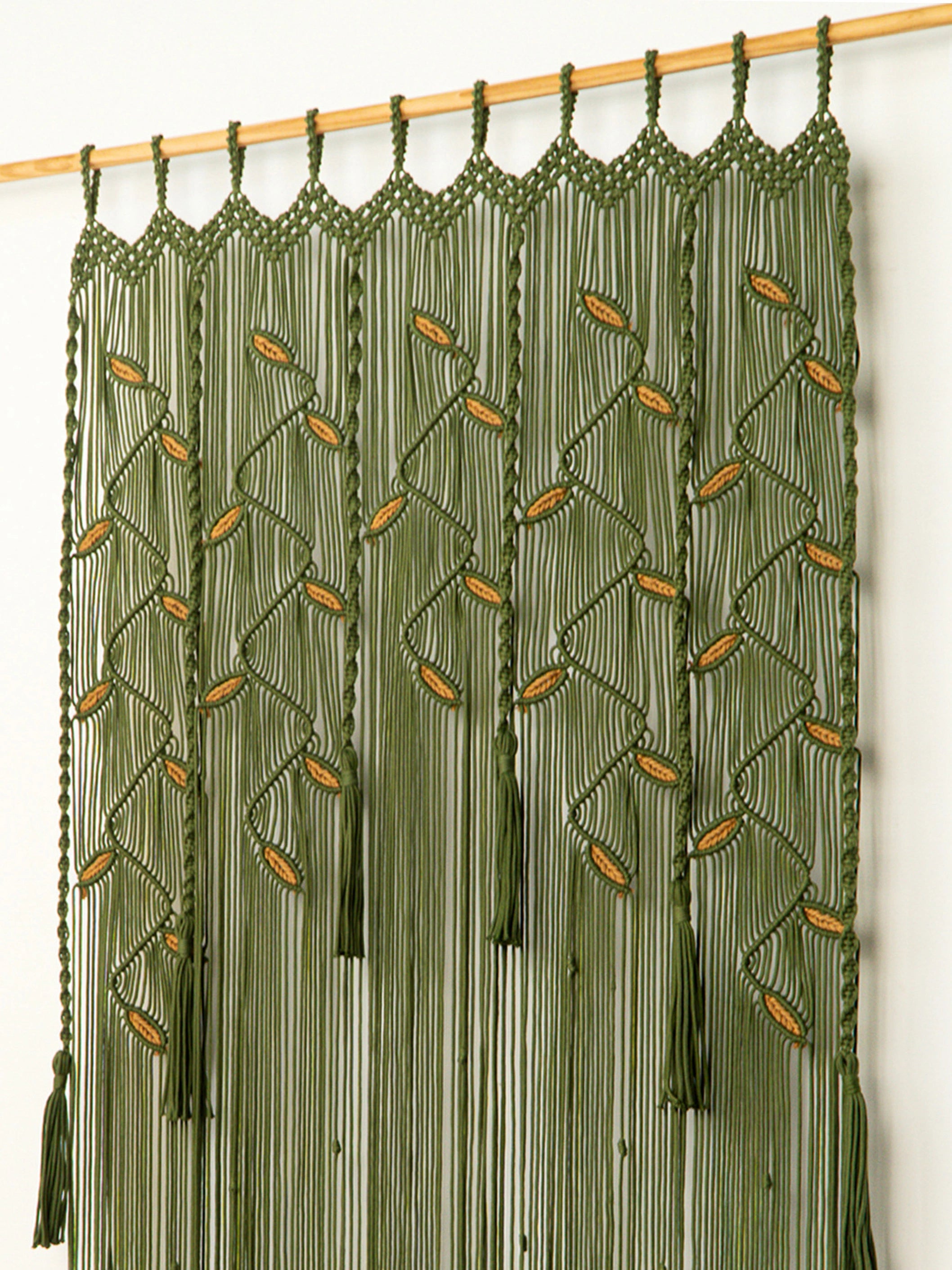 An olive green macrame wall hanging with woven leaf patterns and long fringe, suspended from a wooden rod against a plain white wall.