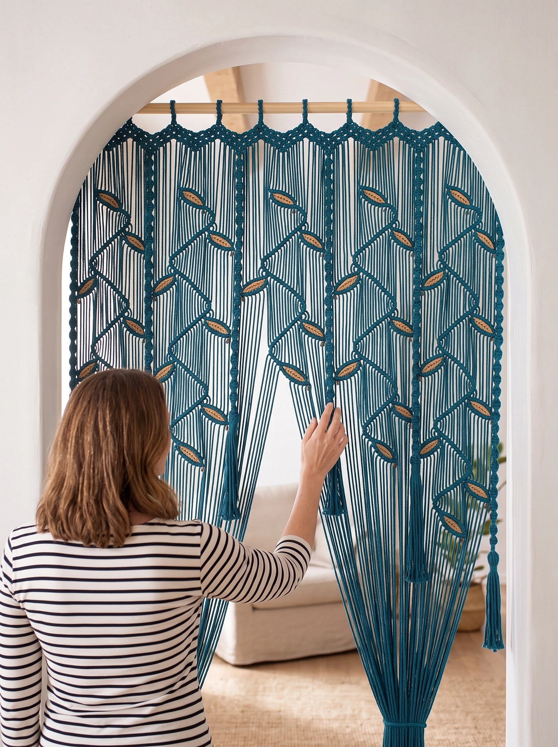 A woman in a black and white striped shirt stands in front of a teal blue macrame curtain with geometric patterns, seen from behind as she touches the hanging textile.