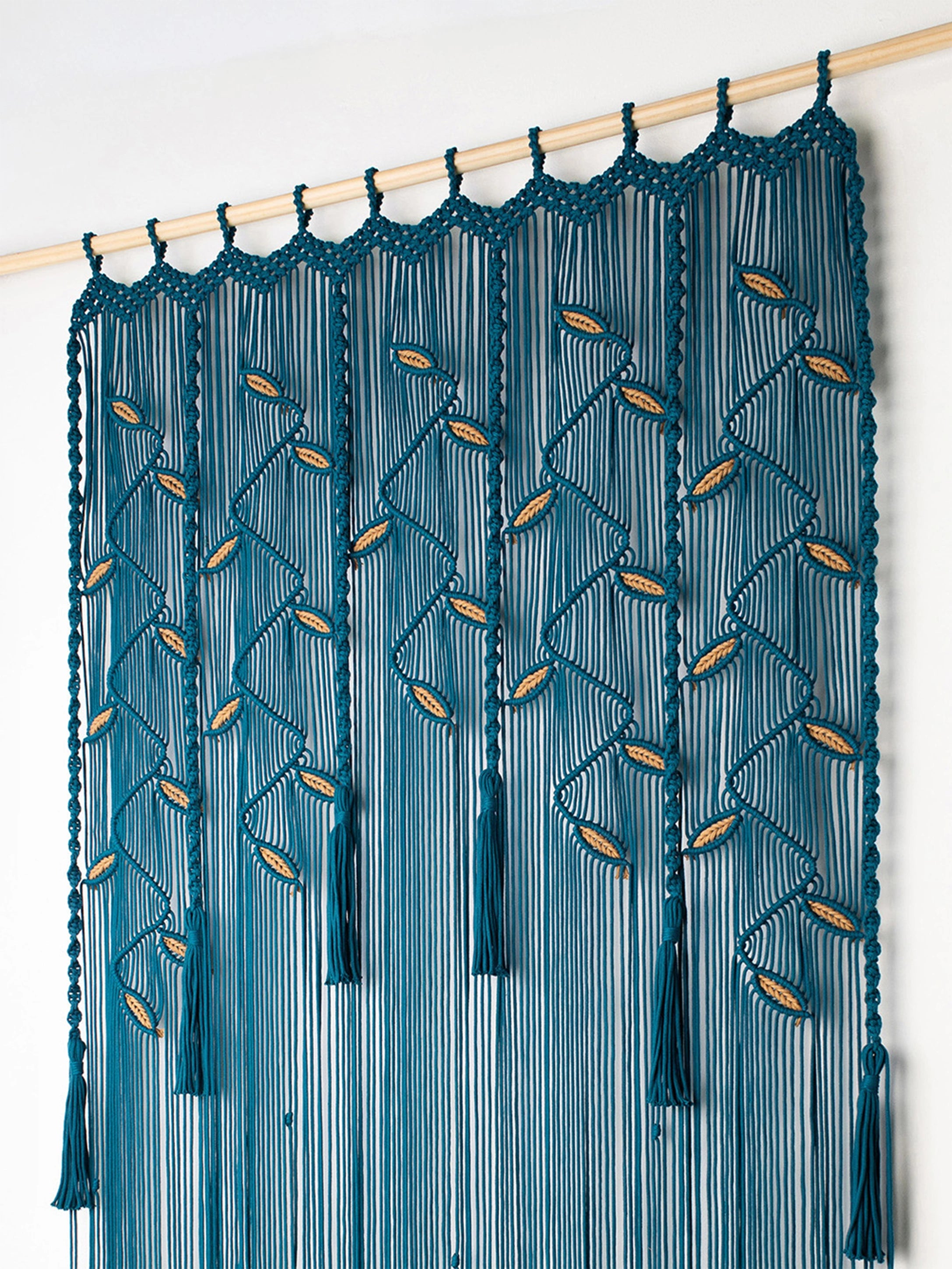 A teal blue macrame wall hanging curtain with intricate woven patterns and tassel ends, suspended from a light wood rod against a plain white wall.