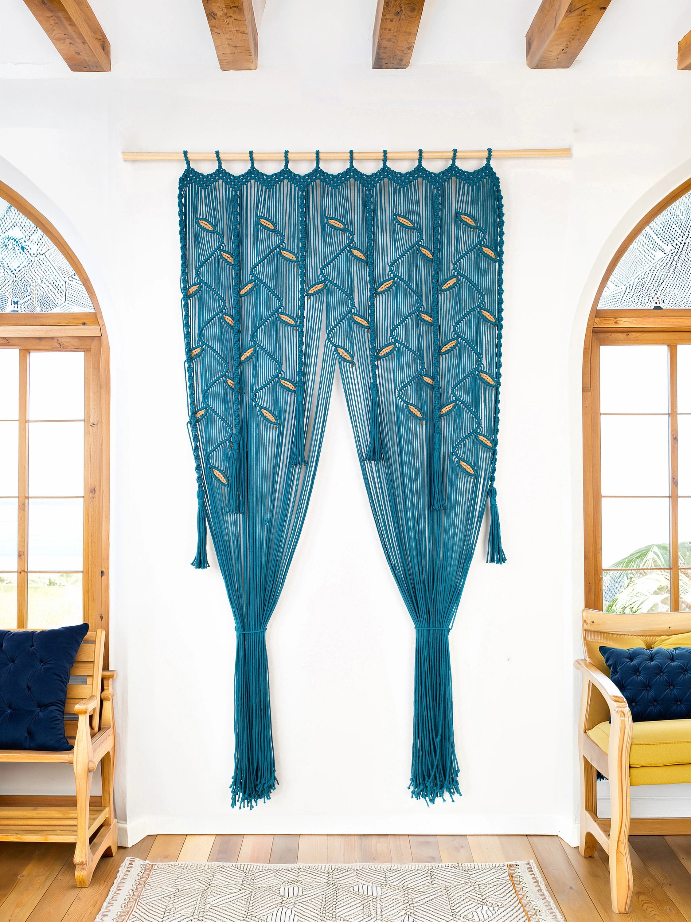 A pair of teal blue sheer curtains with geometric embroidered patterns and tassels, hanging on a wooden rod in a bright room with white walls, wooden ceiling beams, and arched windows.
