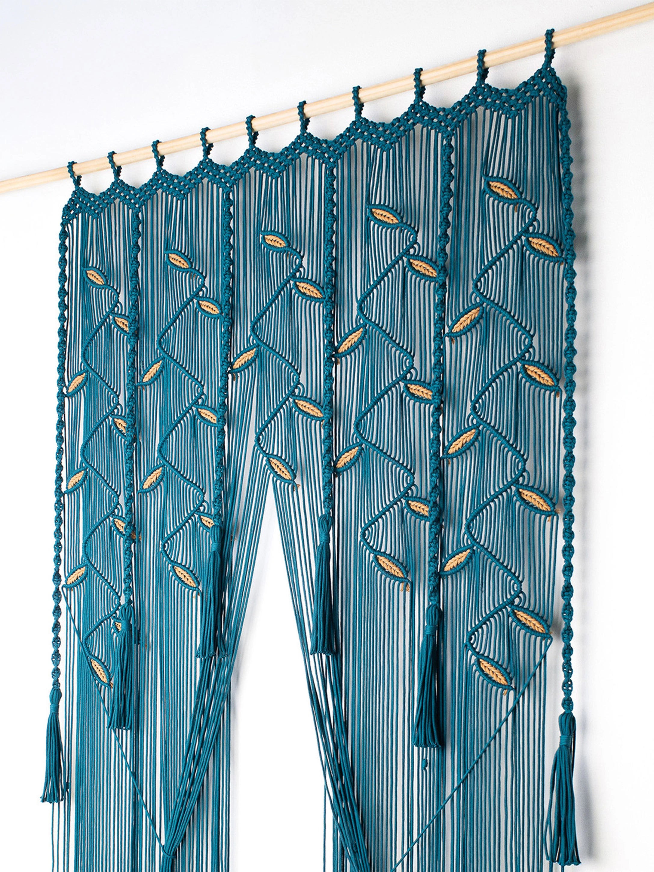 A teal blue macrame curtain panel hanging from a light wood rod against a plain white wall. The curtain features intricate woven patterns with small gold-colored oval accents and long fringe tassels.