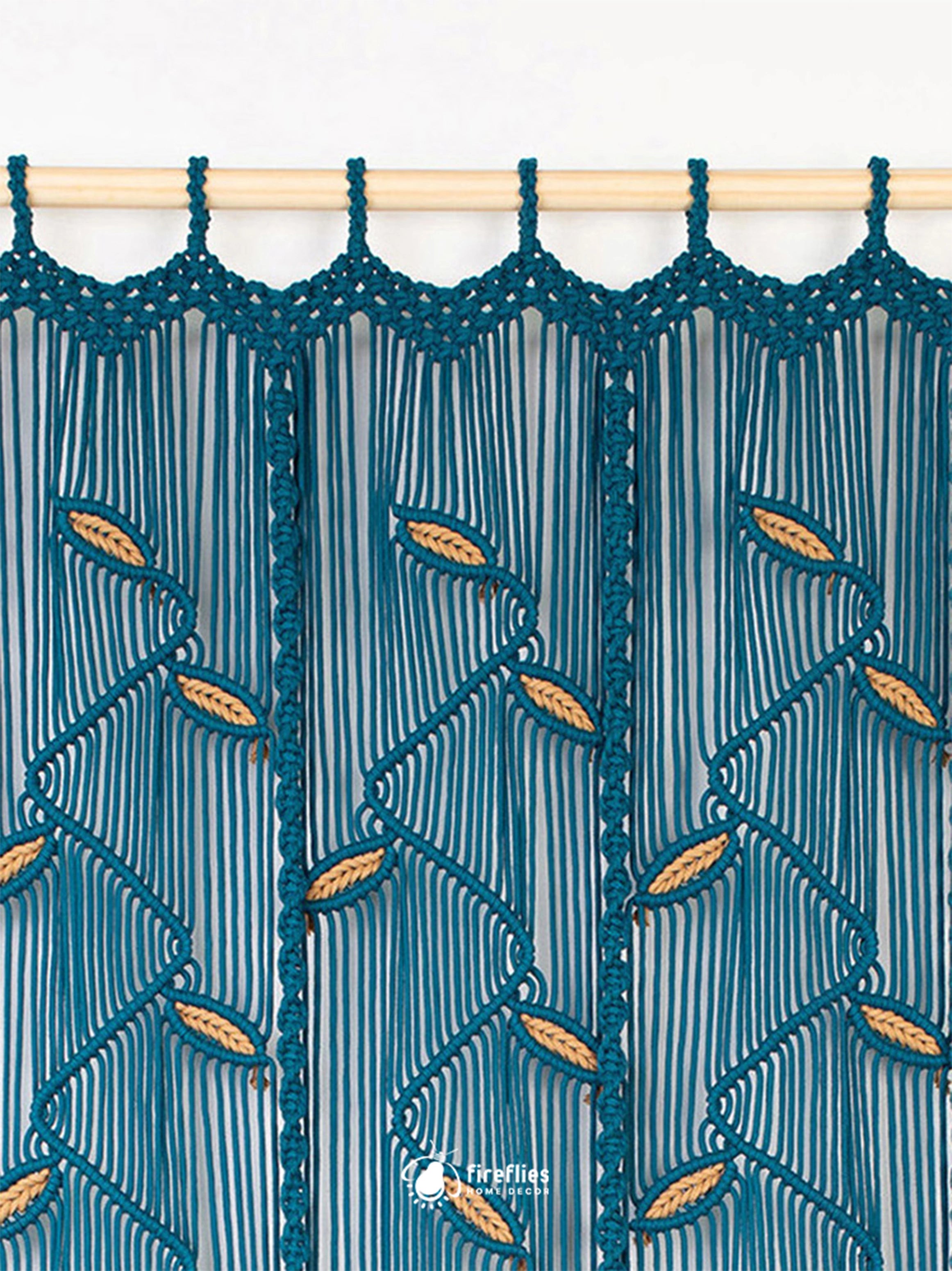 A close-up of a teal macrame curtain panel hanging from a light wood rod against a white wall. The curtain features intricate woven patterns with small tan leaf-like accents and a scalloped top edge.