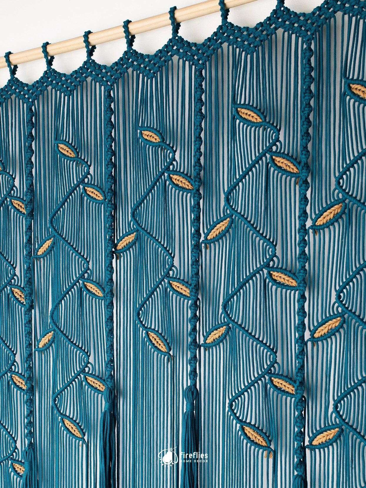 A close-up of a teal blue macrame curtain panel with vertical hanging cords, scalloped top edge, and woven leaf-like accents, shown hanging from a wooden rod against a white wall.