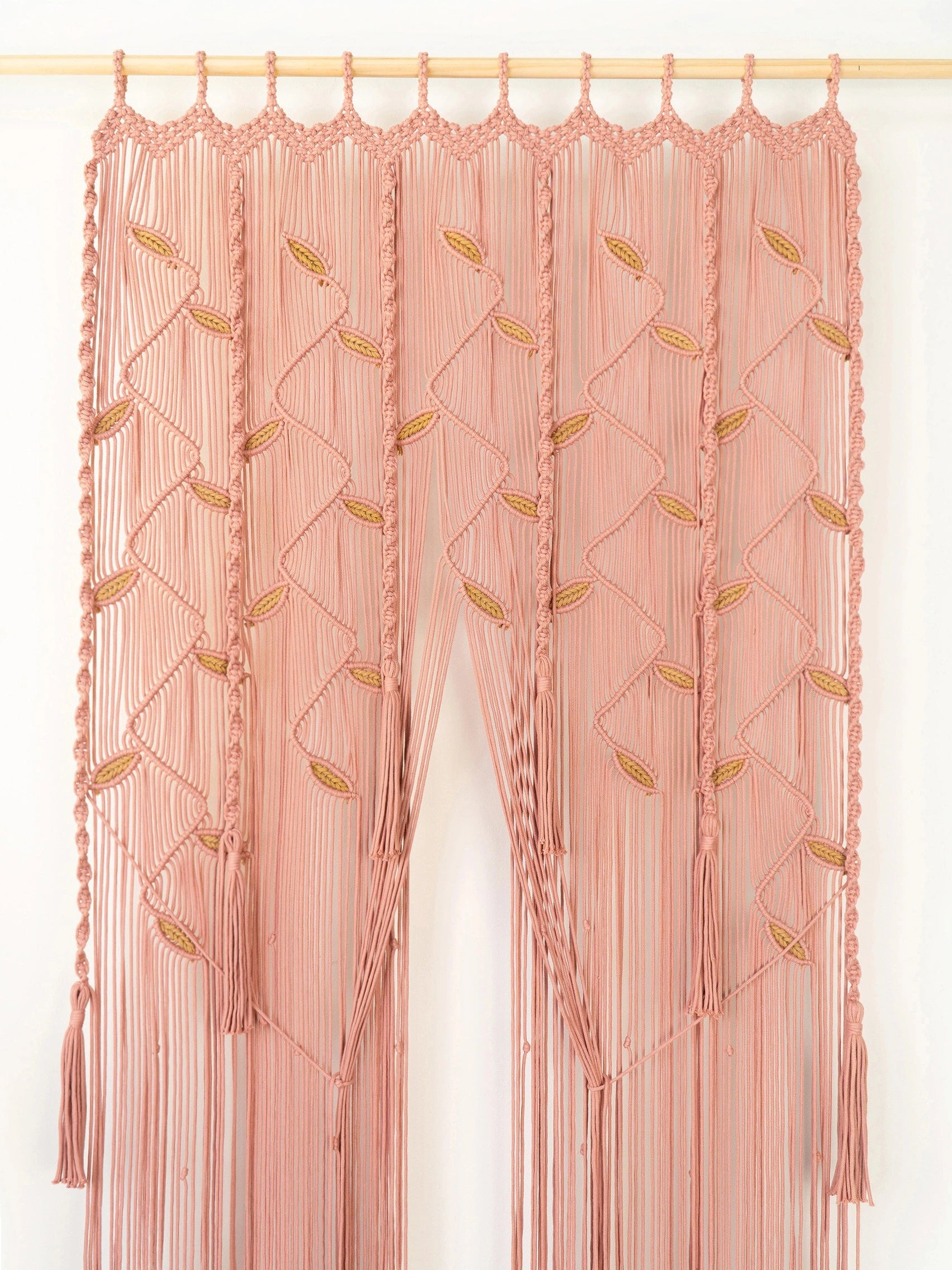 A peach-pink macrame wall hanging curtain with braided tassels and gold leaf embroidery, suspended from a light wood rod against a white wall.