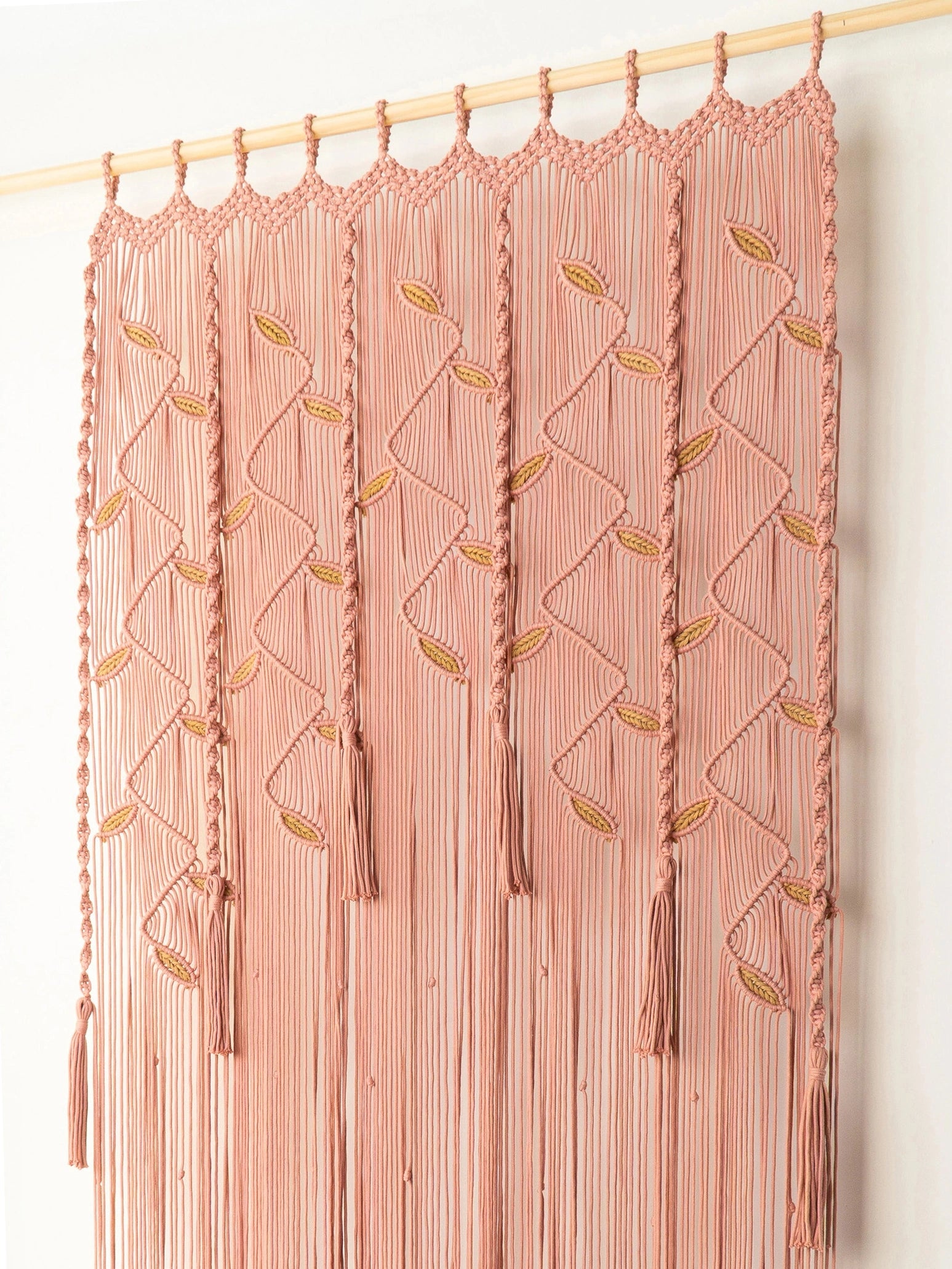 A close-up of a peach-pink fringed curtain or wall hanging with woven geometric diamond patterns and small gold leaf accents.