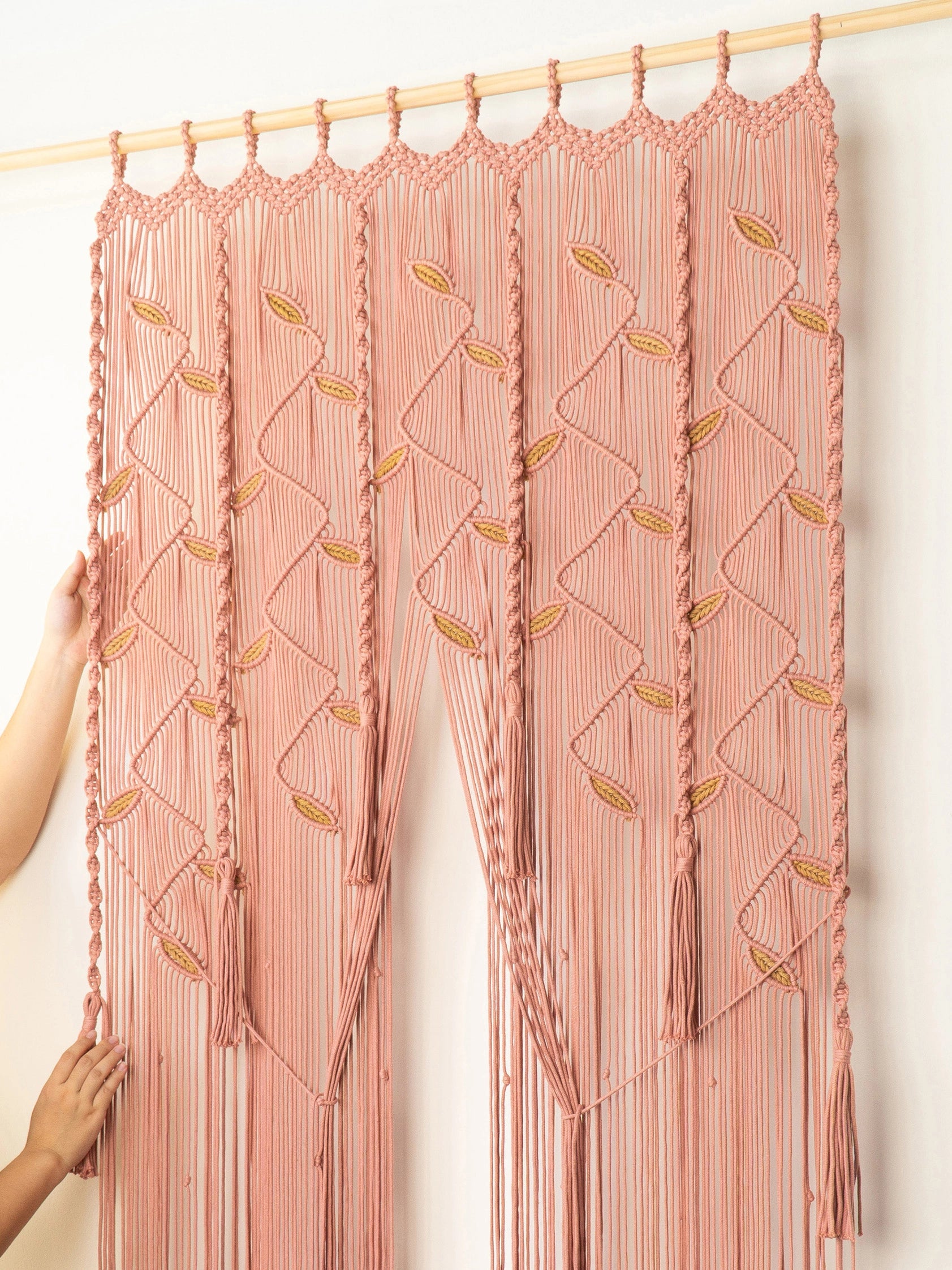 A peach-pink macrame curtain panel hanging from a light wood rod against a plain white wall. The panel features intricate woven geometric patterns, braided vertical strands, and hanging tassels.