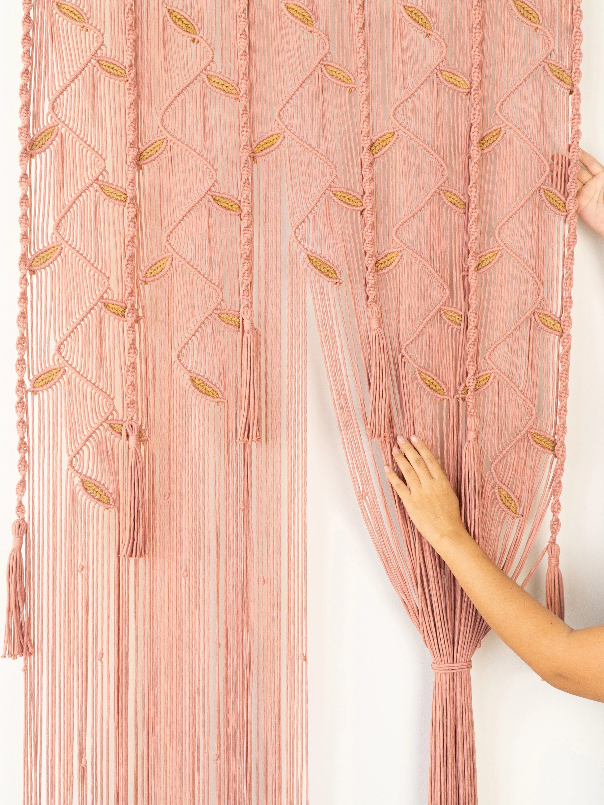A dusty pink macrame curtain panel with geometric woven patterns and long fringe tassels, shown hanging on a white wall with a person's hands adjusting the gathered side.