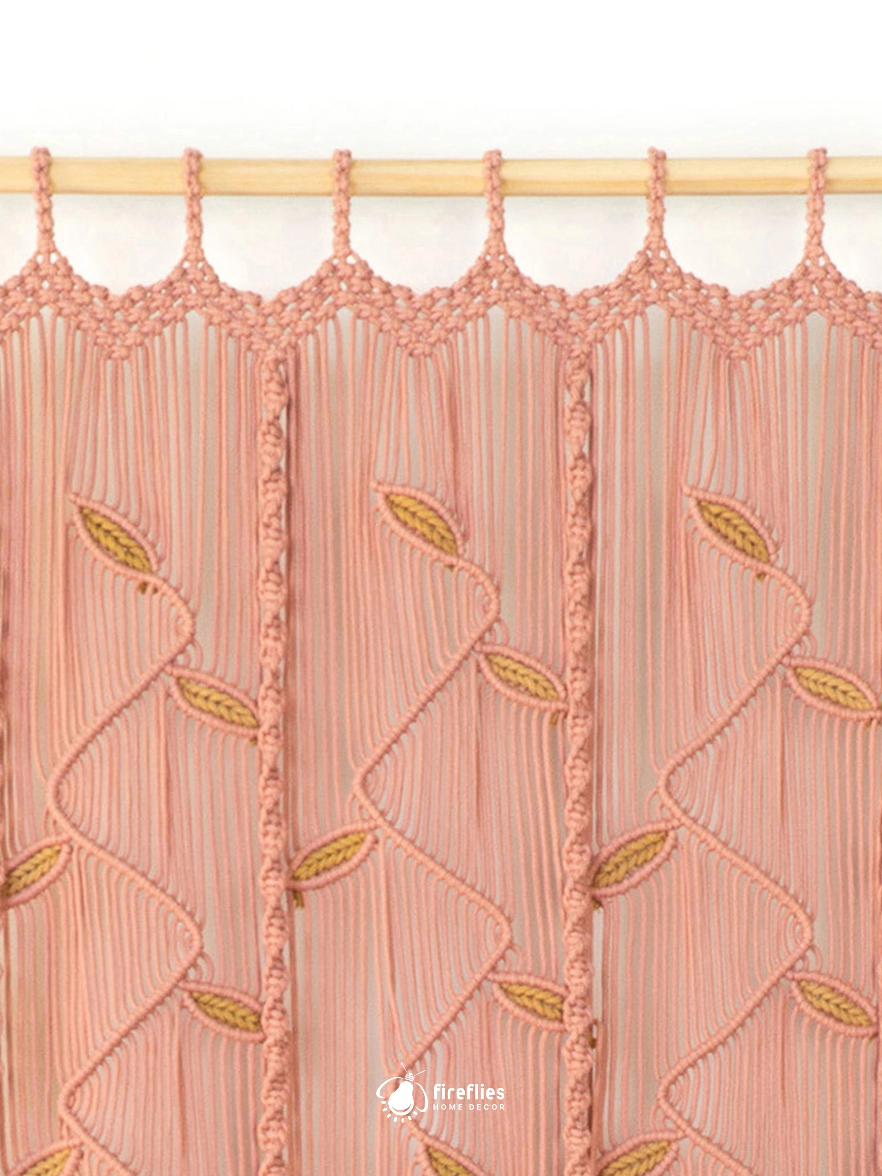 A close-up of a pink macrame curtain panel hanging from a light wood rod. The panel features vertical fringe with woven leaf patterns in olive green and pink.