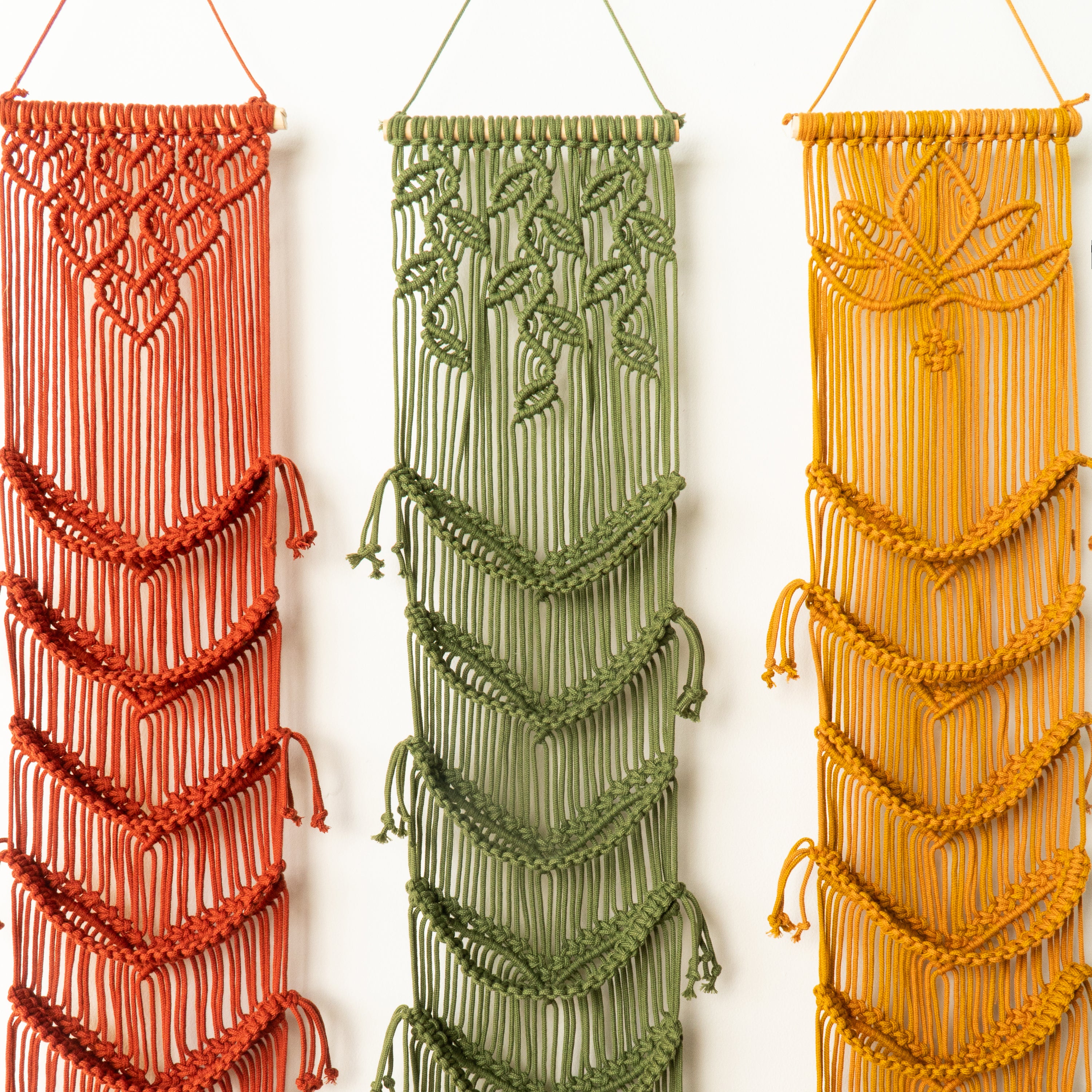Three macrame wall hangings in red, green, and orange on a white background