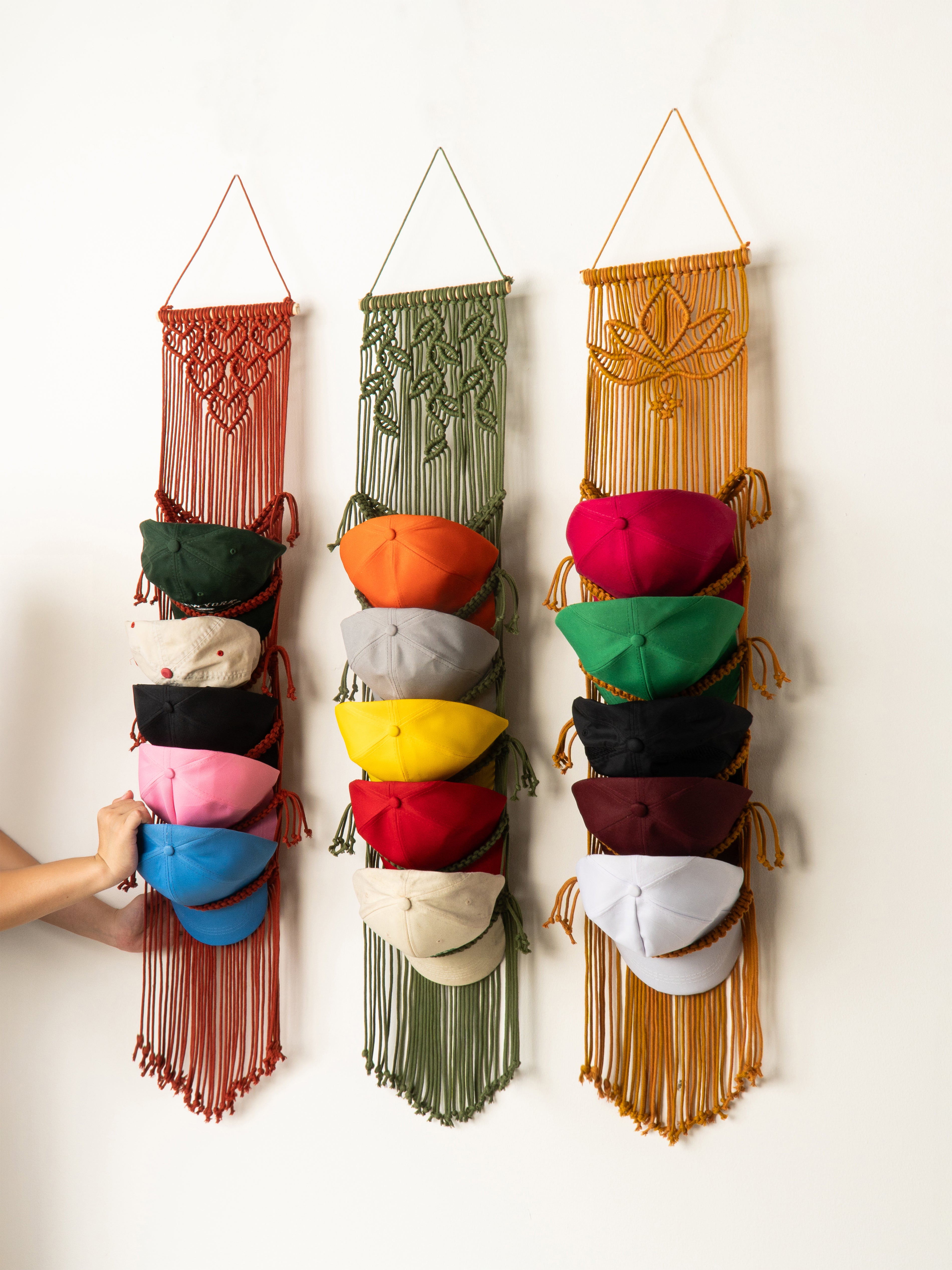 Three colorful macrame hat racks with hats on a white wall.