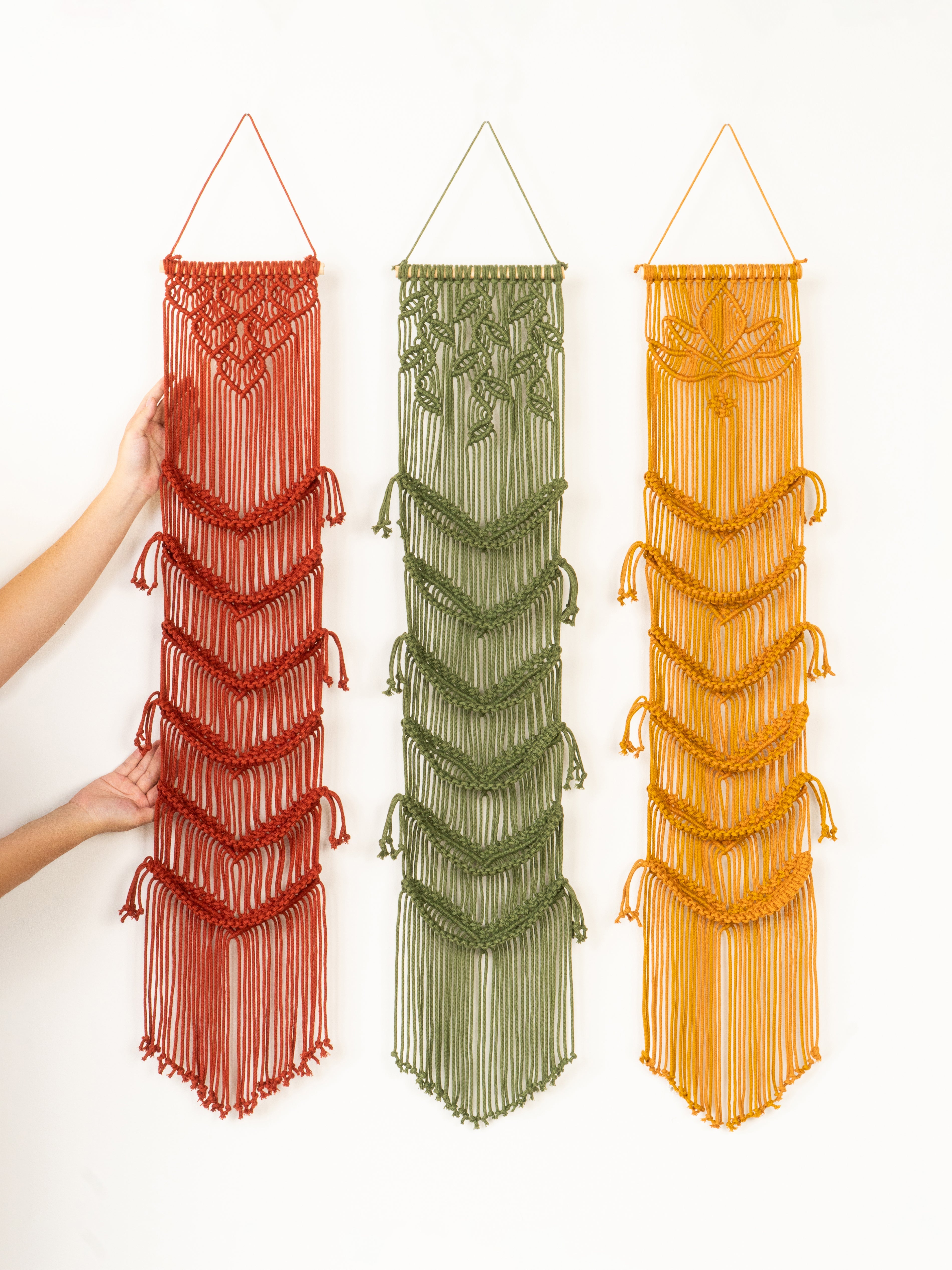 Three macrame wall hangings in red, green, and orange on a white background.