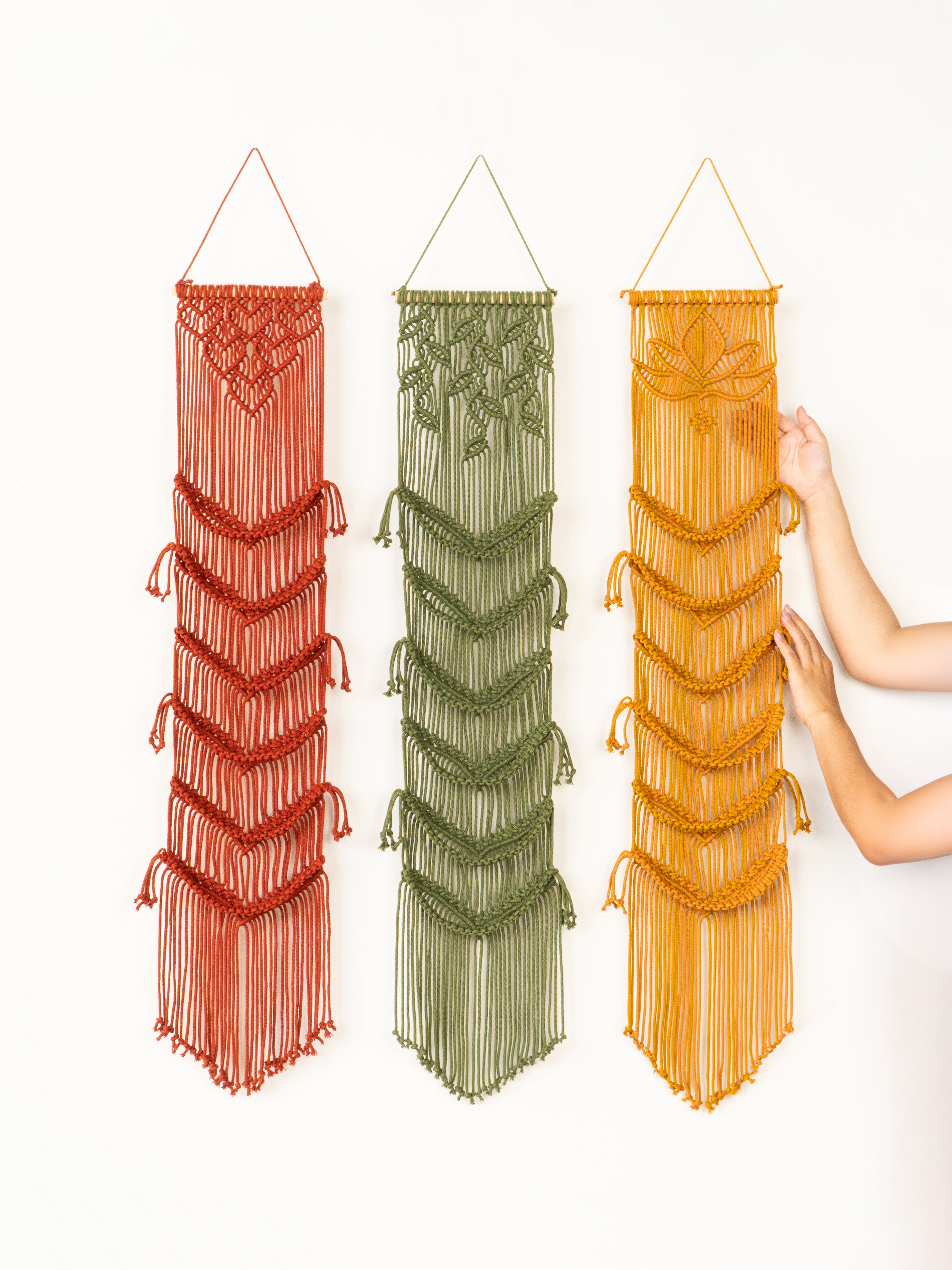 Three macrame wall hangings in red, green, and orange on a white background.