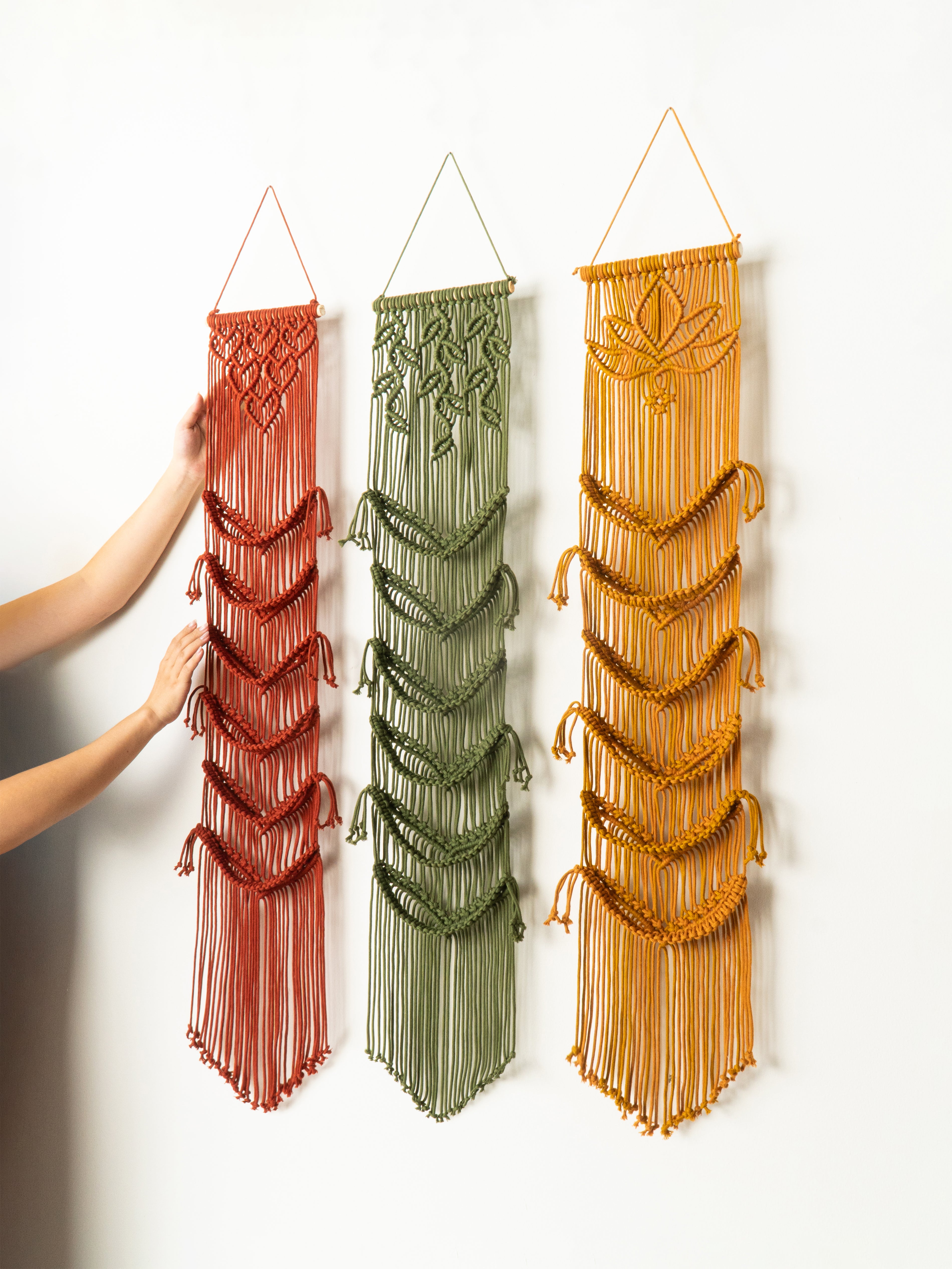 Three macrame wall hangings in red, green, and orange hanging on a white wall.
