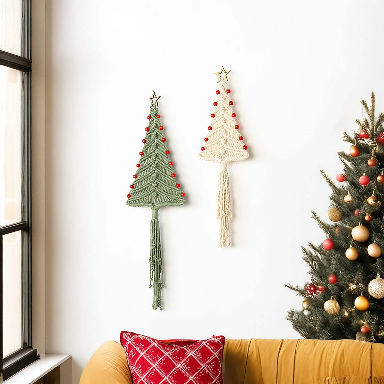Two macrame Christmas tree wall hangings on a white wall next to a decorated Christmas tree.