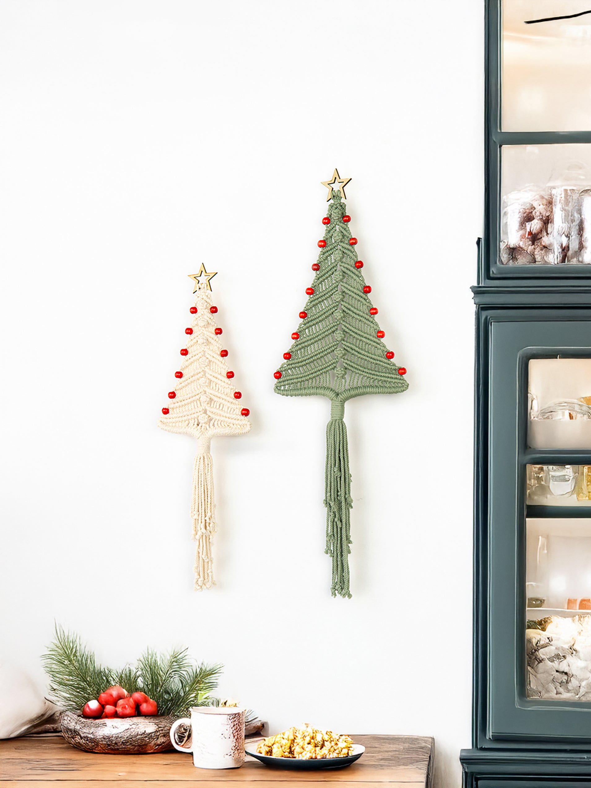 Two macrame Christmas tree wall hangings on a white wall with a cabinet and decorative items in the background.