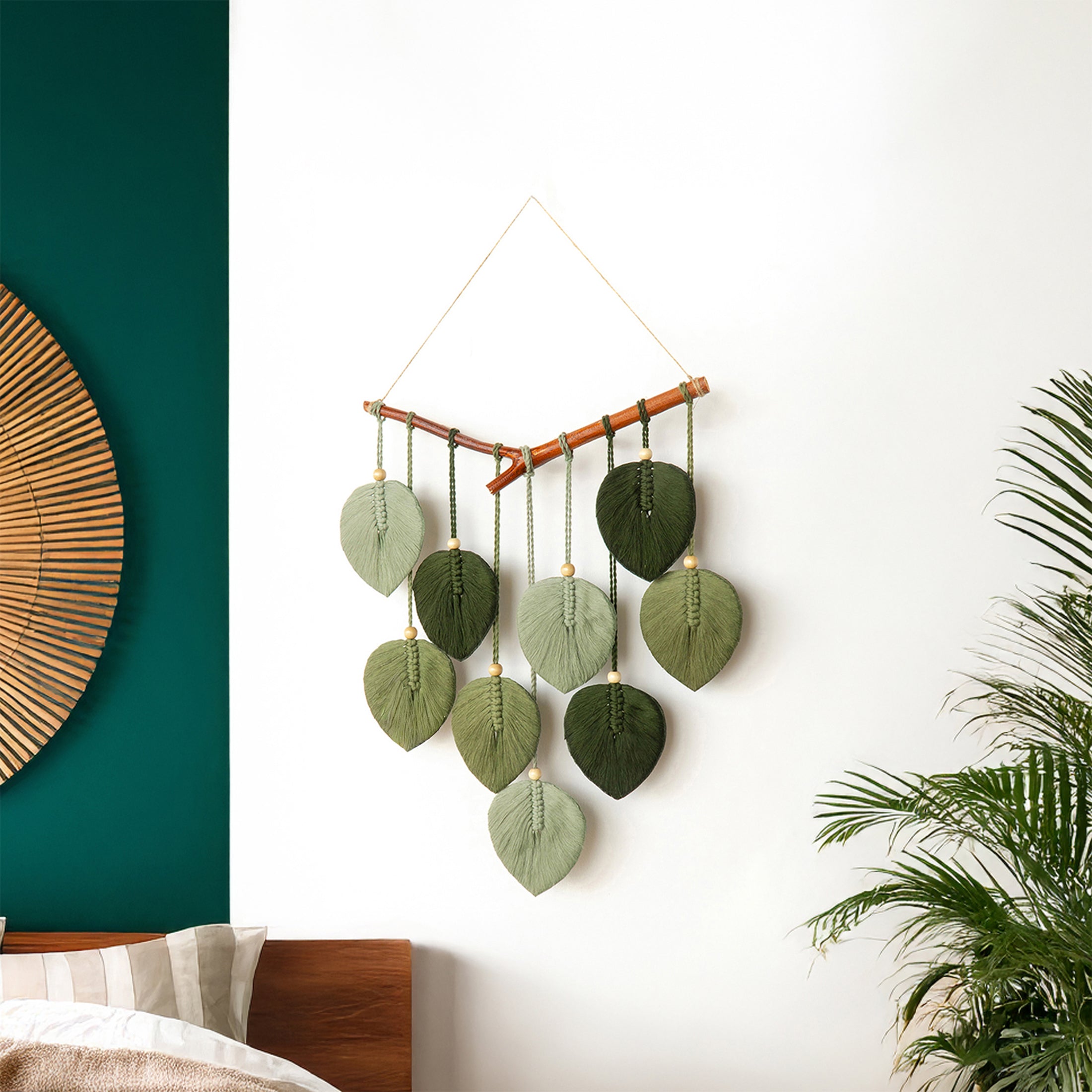 Decorative wall hanging with green leaves on a white wall next to a bed with a wooden headboard.