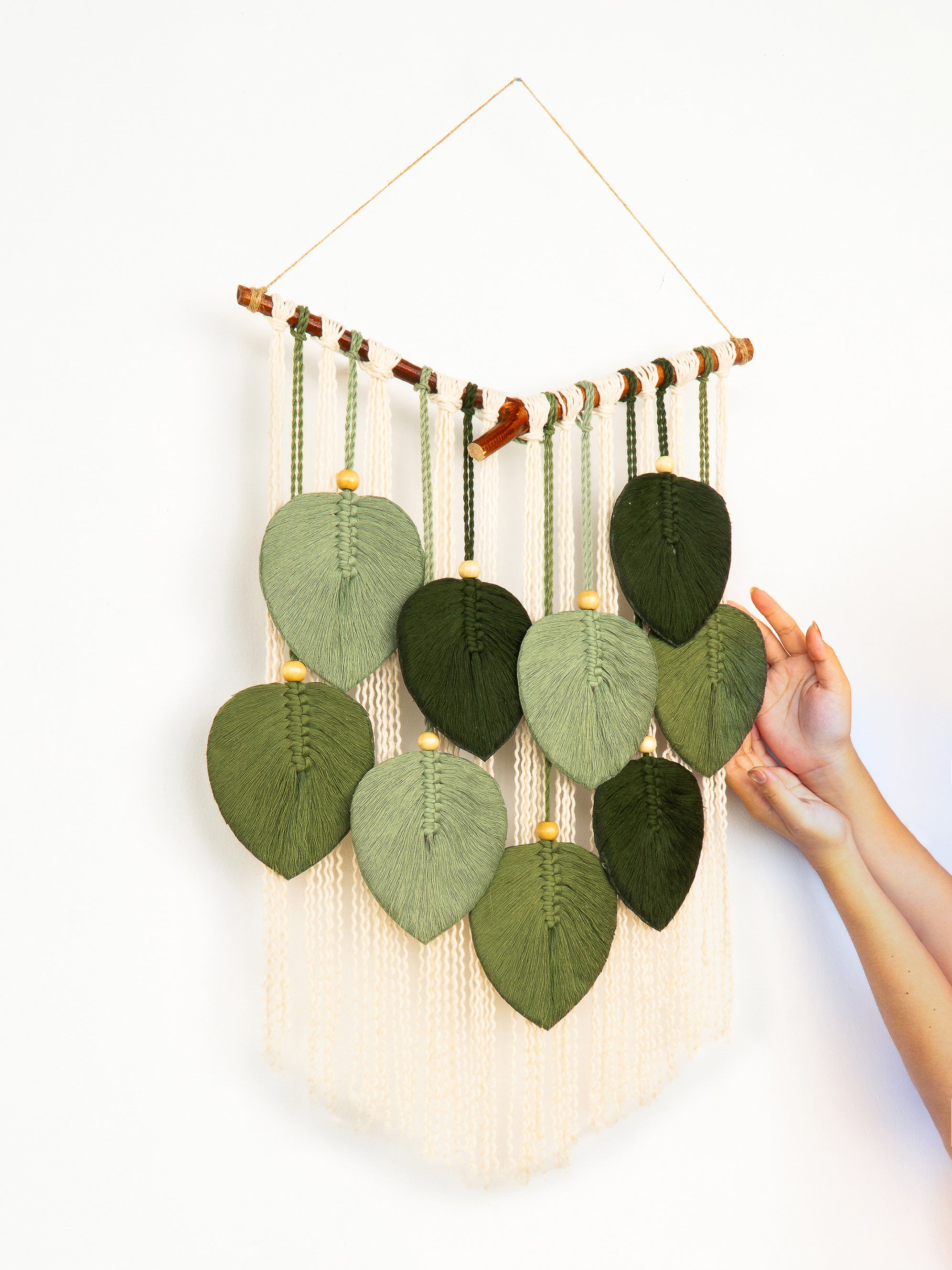 Handmade wall hanging with knitted leaf patterns and macrame strings, held by a person.