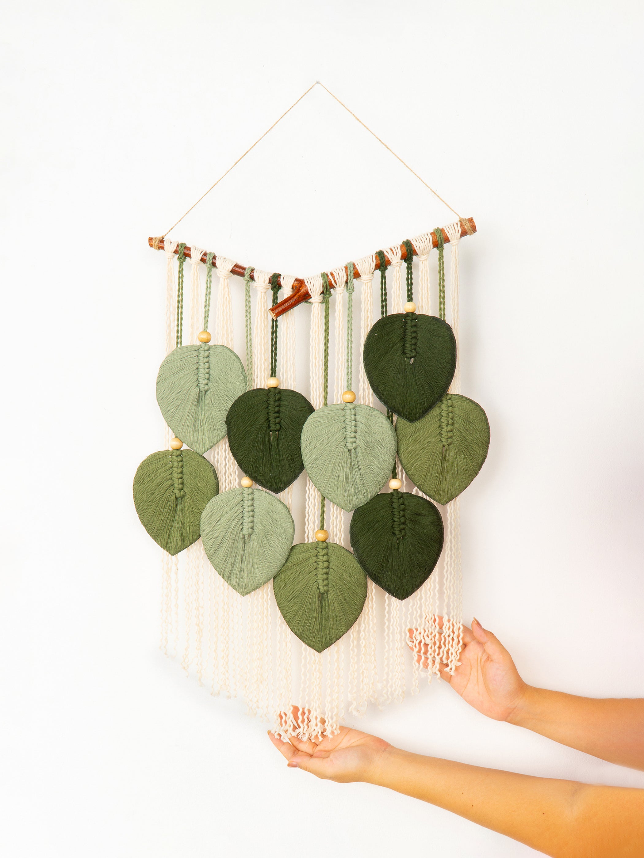 Handmade wall hanging with green leaf-shaped fabric pieces and braided strings on a white background.
