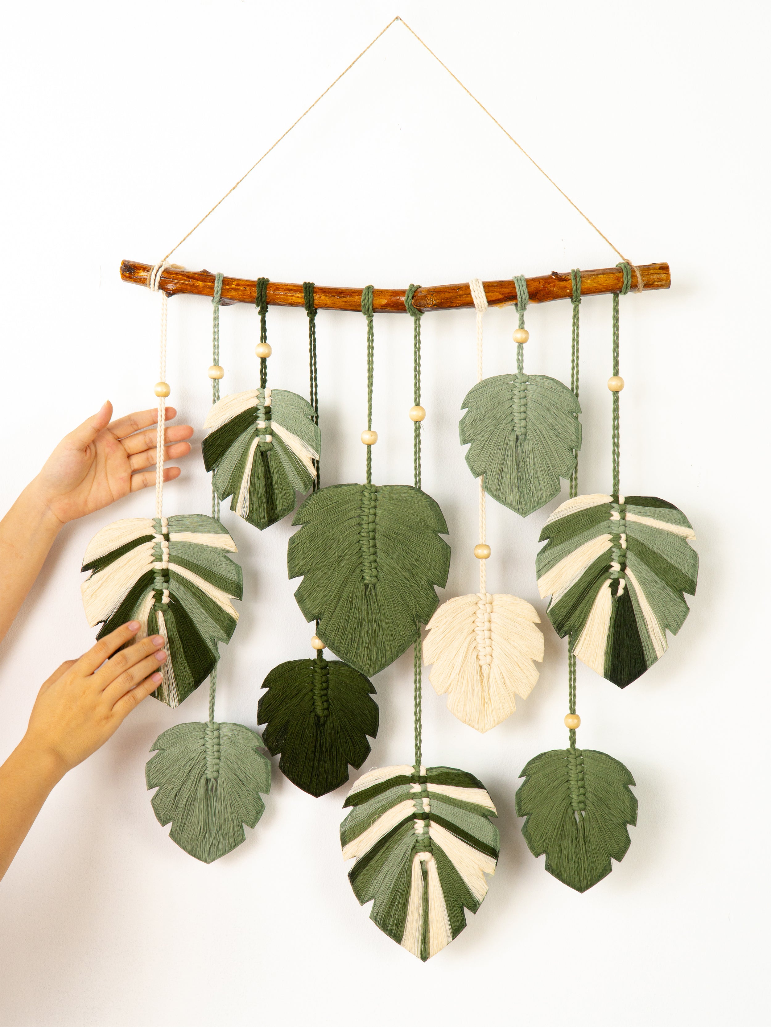 Handmade wall hanging with green leaf-shaped fabric pieces and braided strings on a white background.