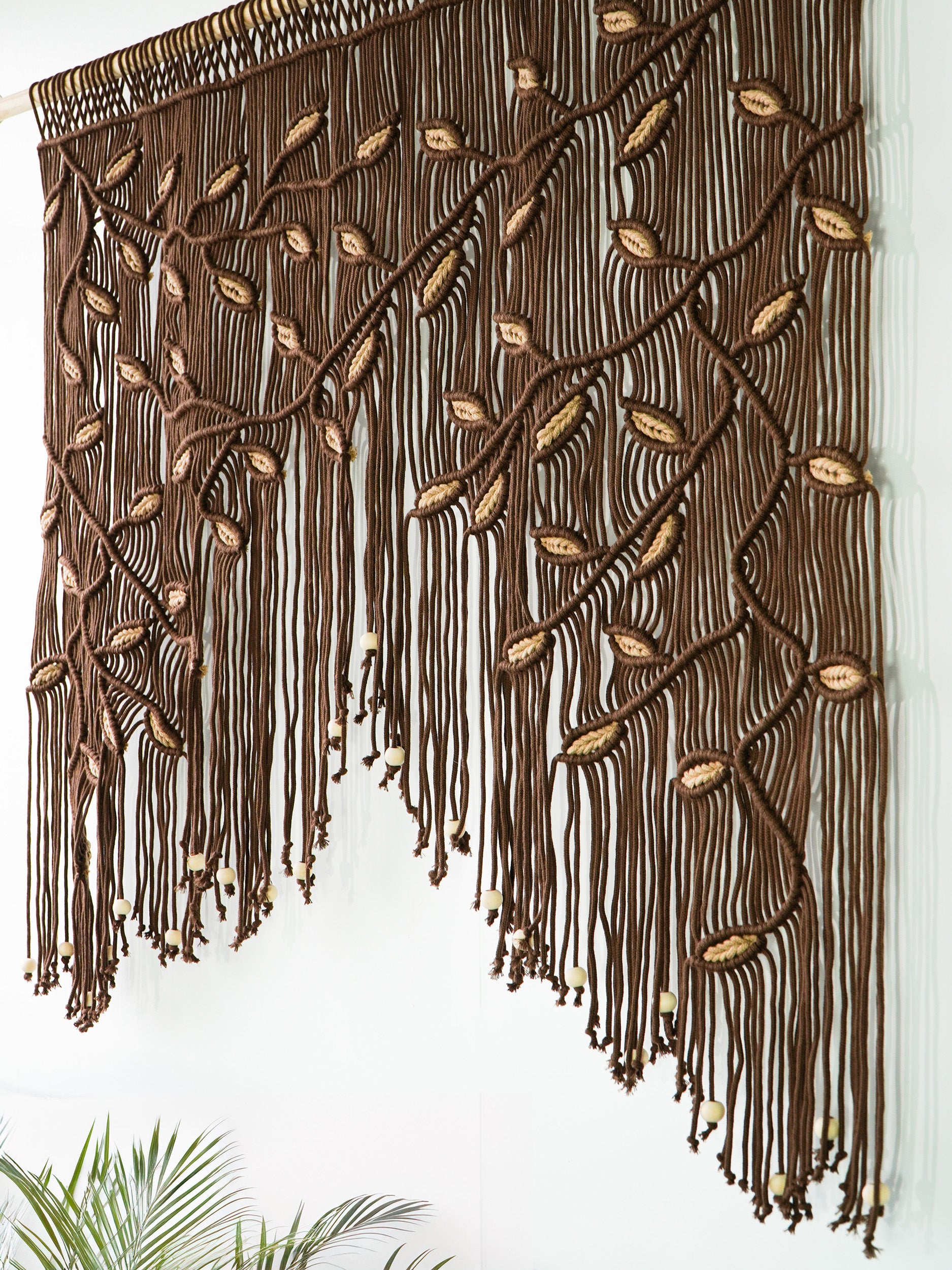 Artisan fiber art in deep brown with meticulous wheat-colored leaf knotting on a natural wood dowel.