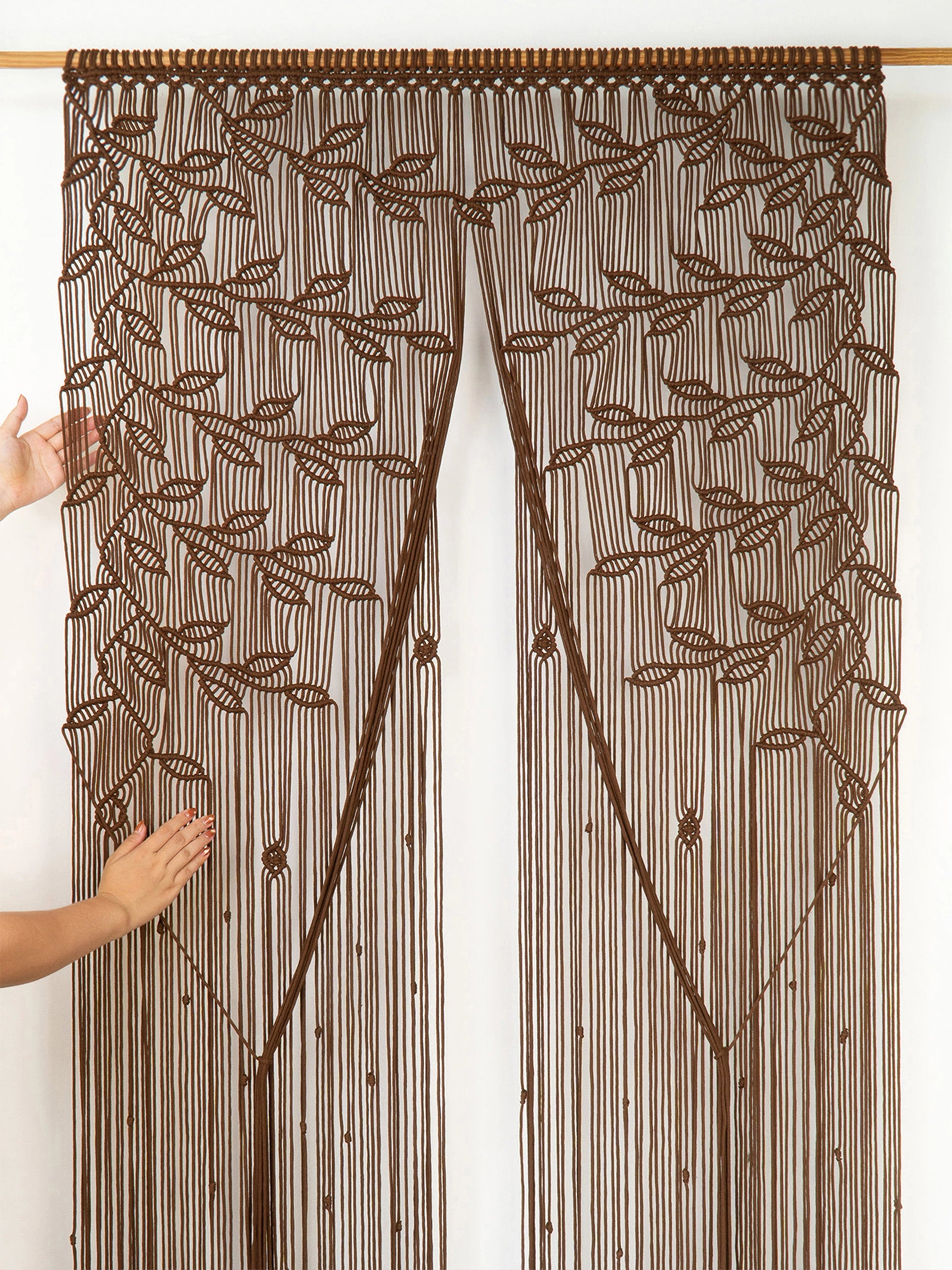A brown beaded curtain with intricate leaf and vine patterns, hanging from a wooden rod. Two hands are visible on the left side holding the curtain open to show its full length and texture.