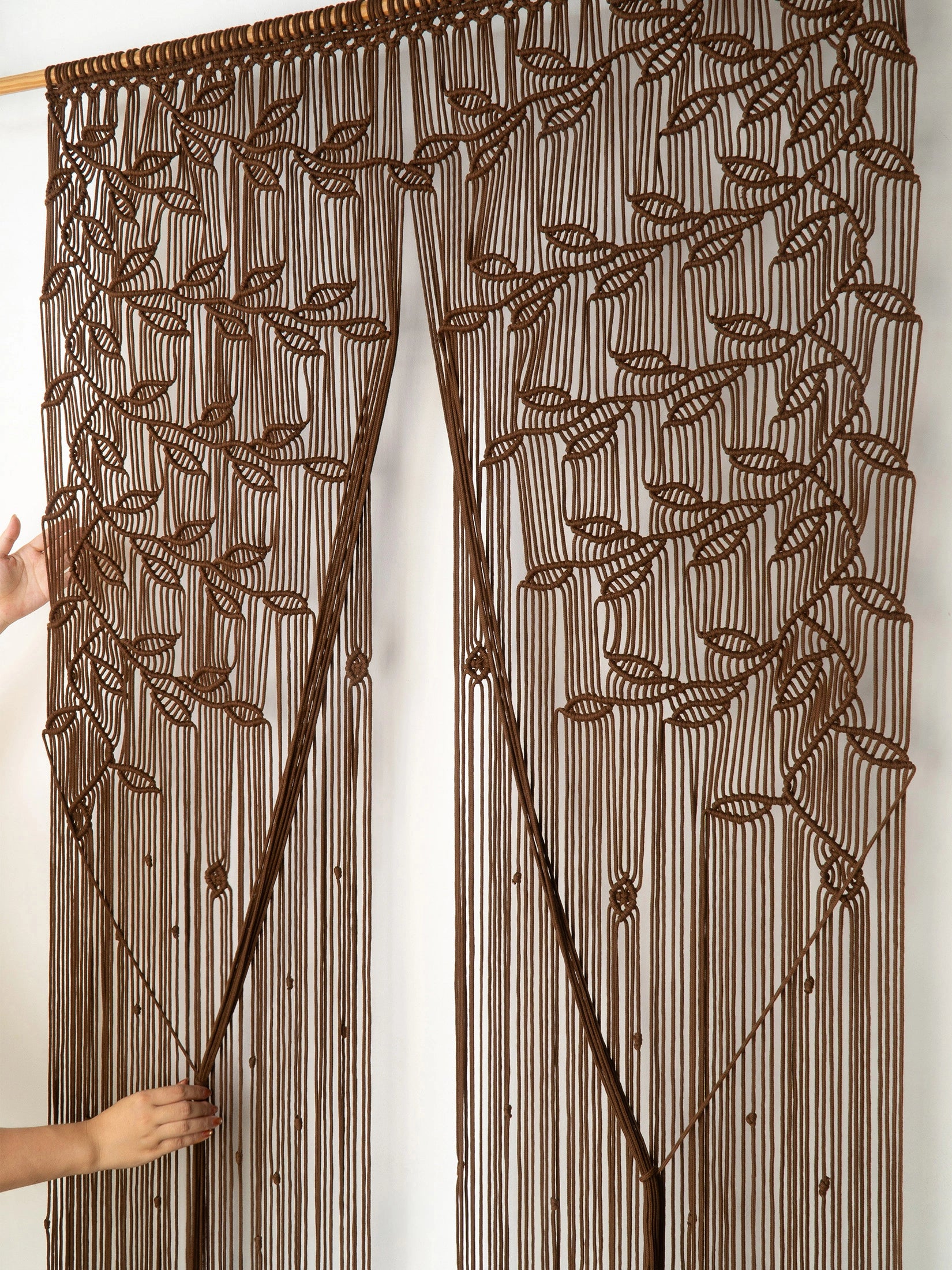 A brown beaded curtain with an embroidered leaf pattern, held open by two hands against a plain white background.