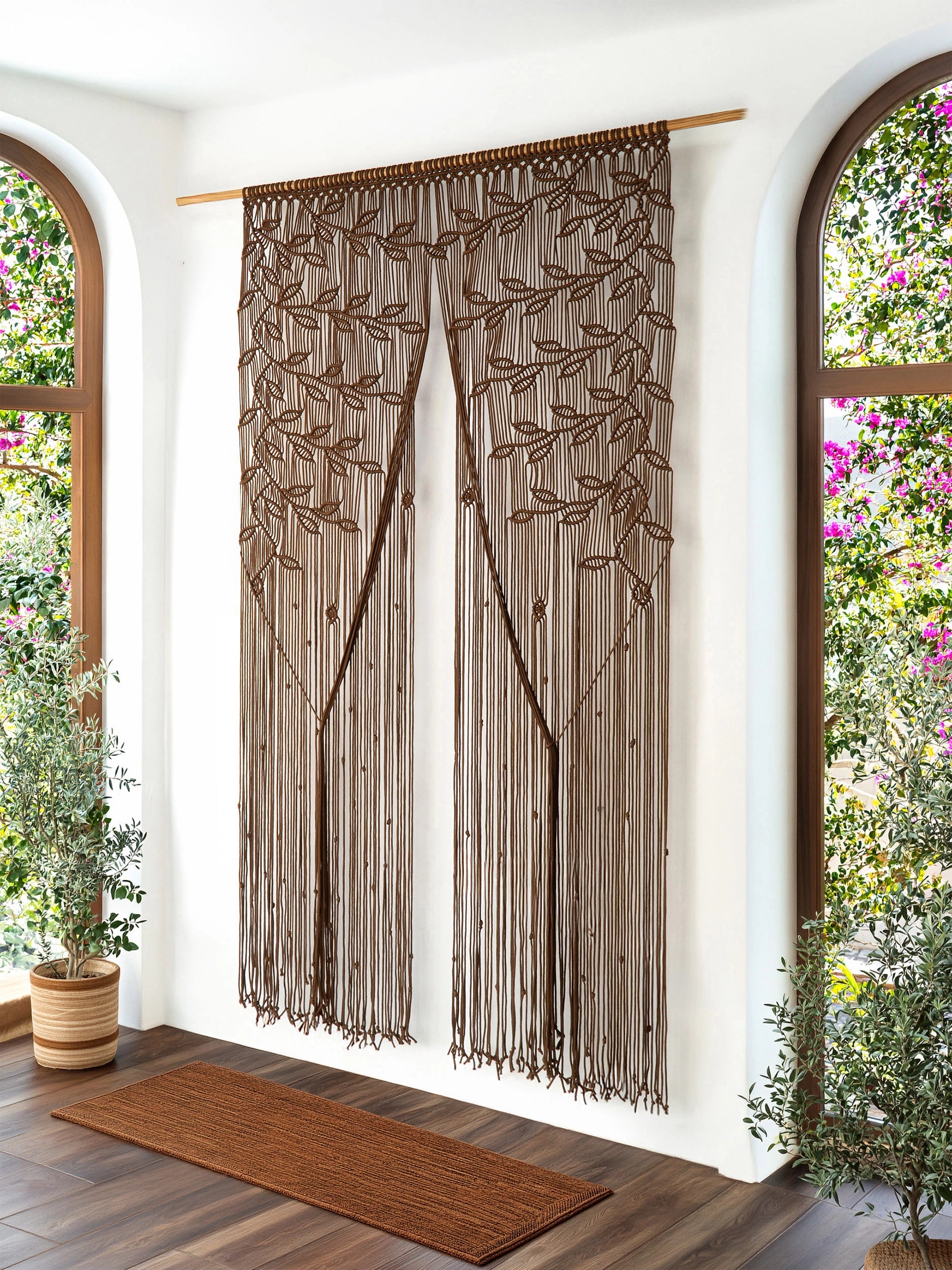 A pair of brown curtain panels with an embroidered tree design and long fringe, hanging on a wooden rod in a bright room with arched windows and plants.
