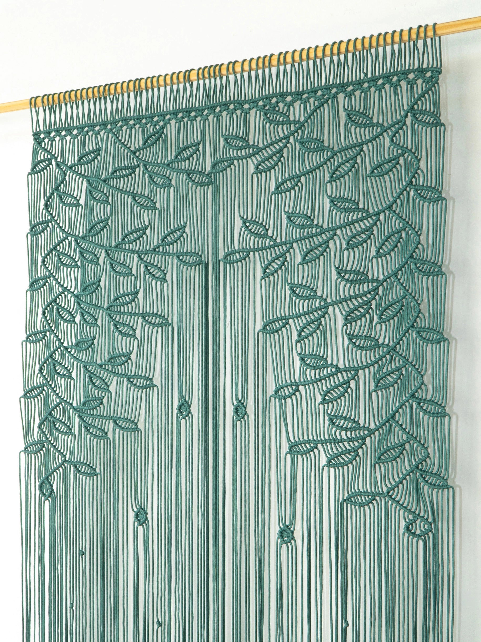 A green embroidered curtain panel hanging from a gold rod against a plain white wall, featuring vertical fringe with leaf and vine embroidery.