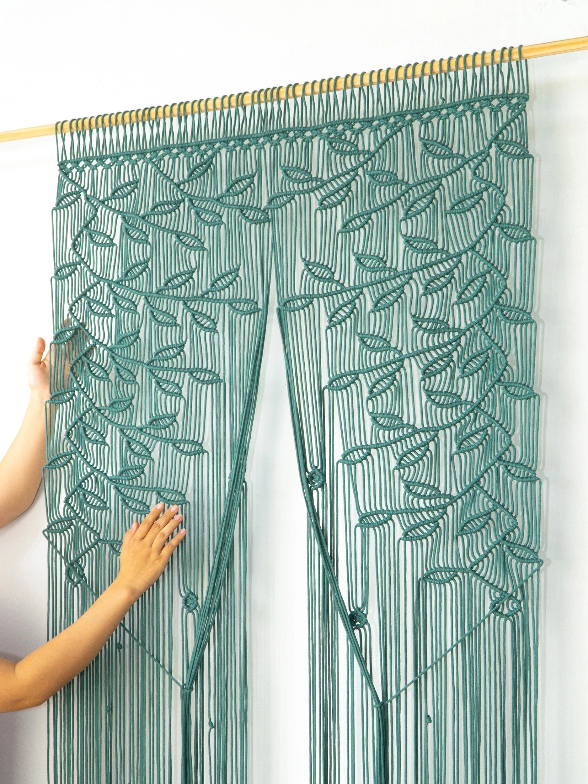 A green macrame curtain with a leafy vine pattern hanging from a wooden rod, with a person's hands adjusting the left panel against a plain white wall.