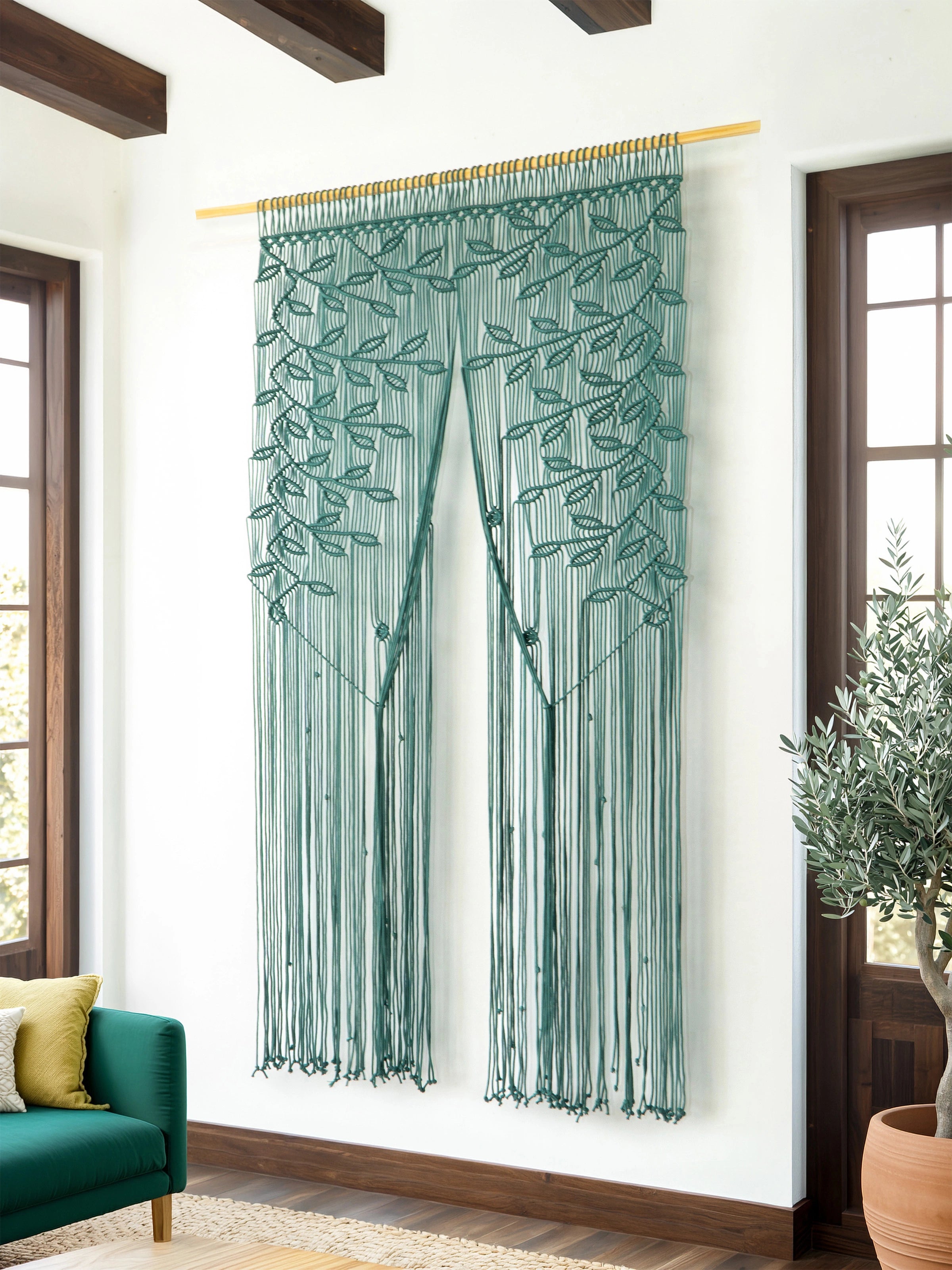 A teal green beaded curtain with embroidered leafy vine patterns hangs from a gold rod in a bright room with white walls, dark wood ceiling beams, and a potted olive tree nearby.