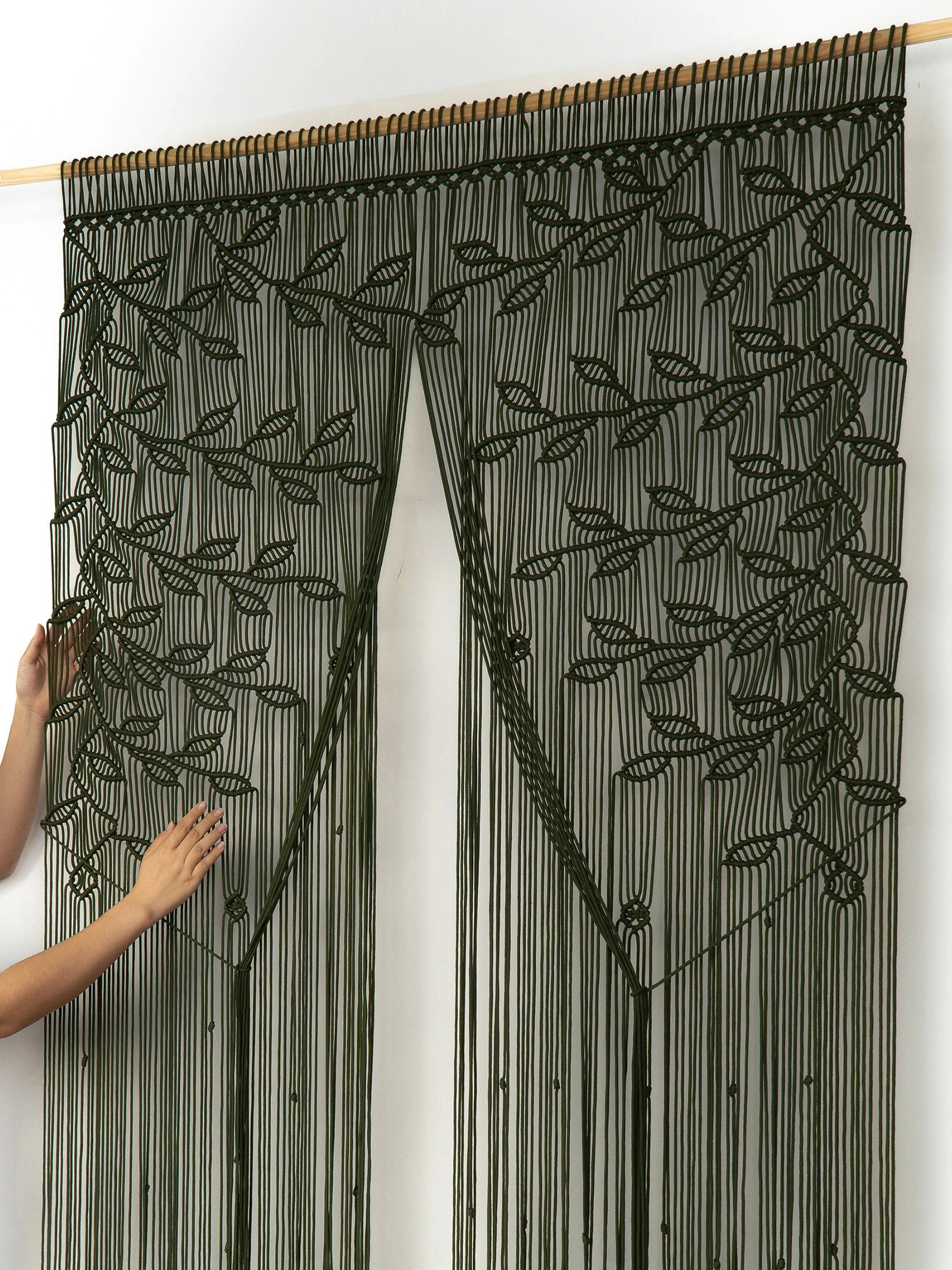A dark olive green beaded curtain with a leafy vine pattern hanging from a wooden rod, with a person's hands visible on the left side adjusting or displaying the fabric.