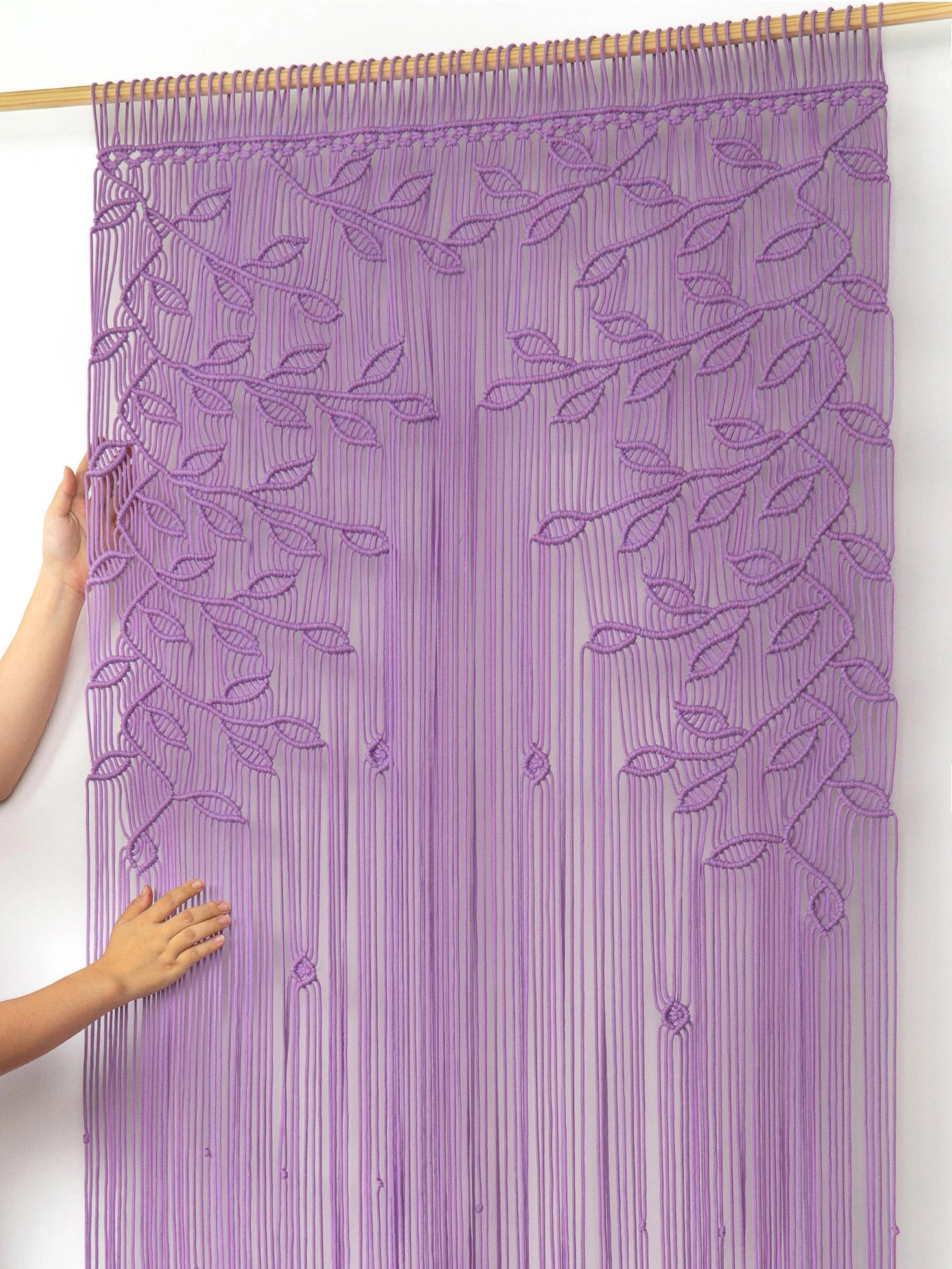 A purple embroidered curtain panel hanging from a wooden rod, with a person's hands visible holding the left side to show its drape against a plain white wall.