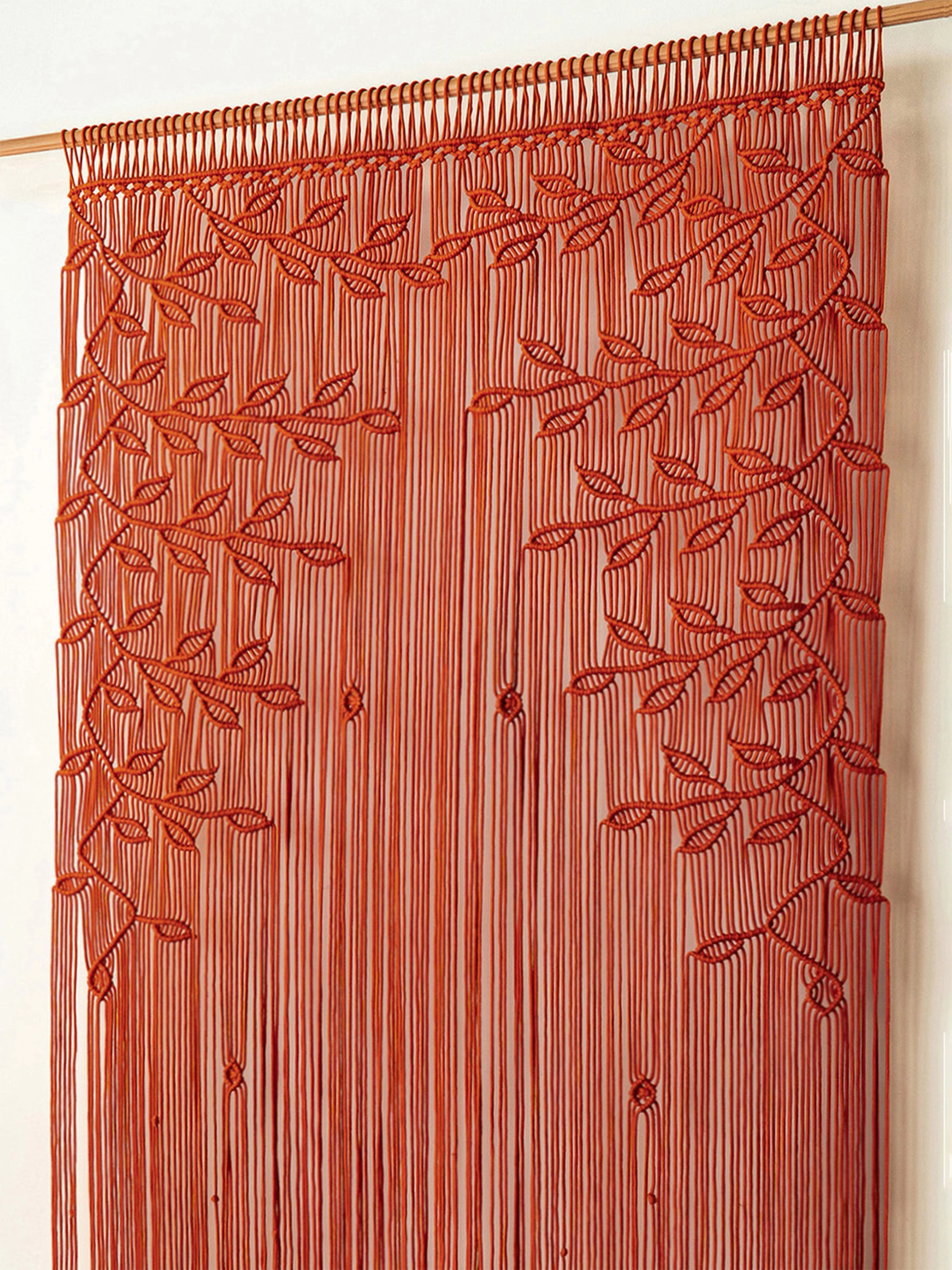 A rust-orange beaded curtain featuring vertical strands with leafy vine patterns, hanging from a wooden rod against a plain off-white wall.
