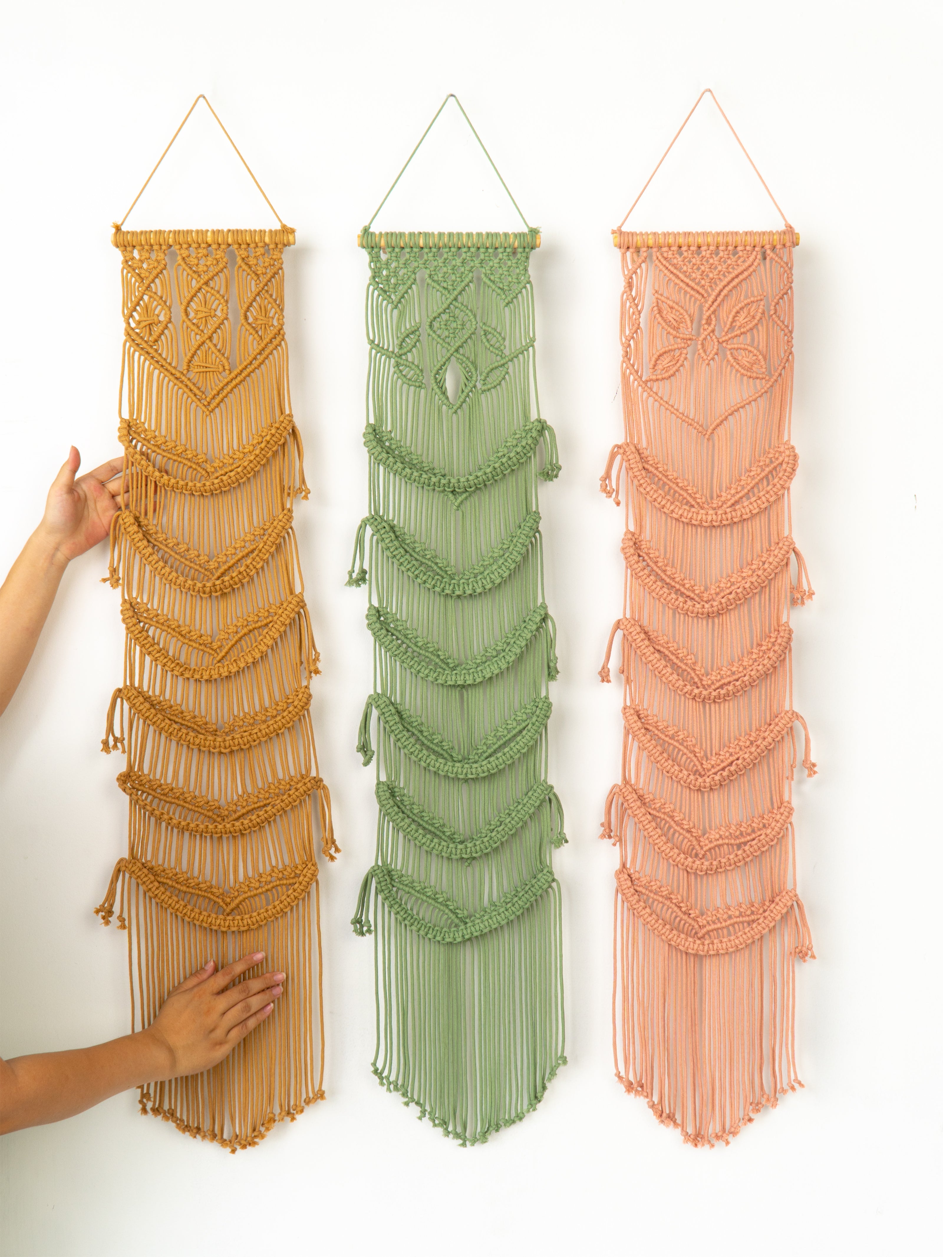 Three macrame wall hangings in gold, green, and peach colors on a white background.