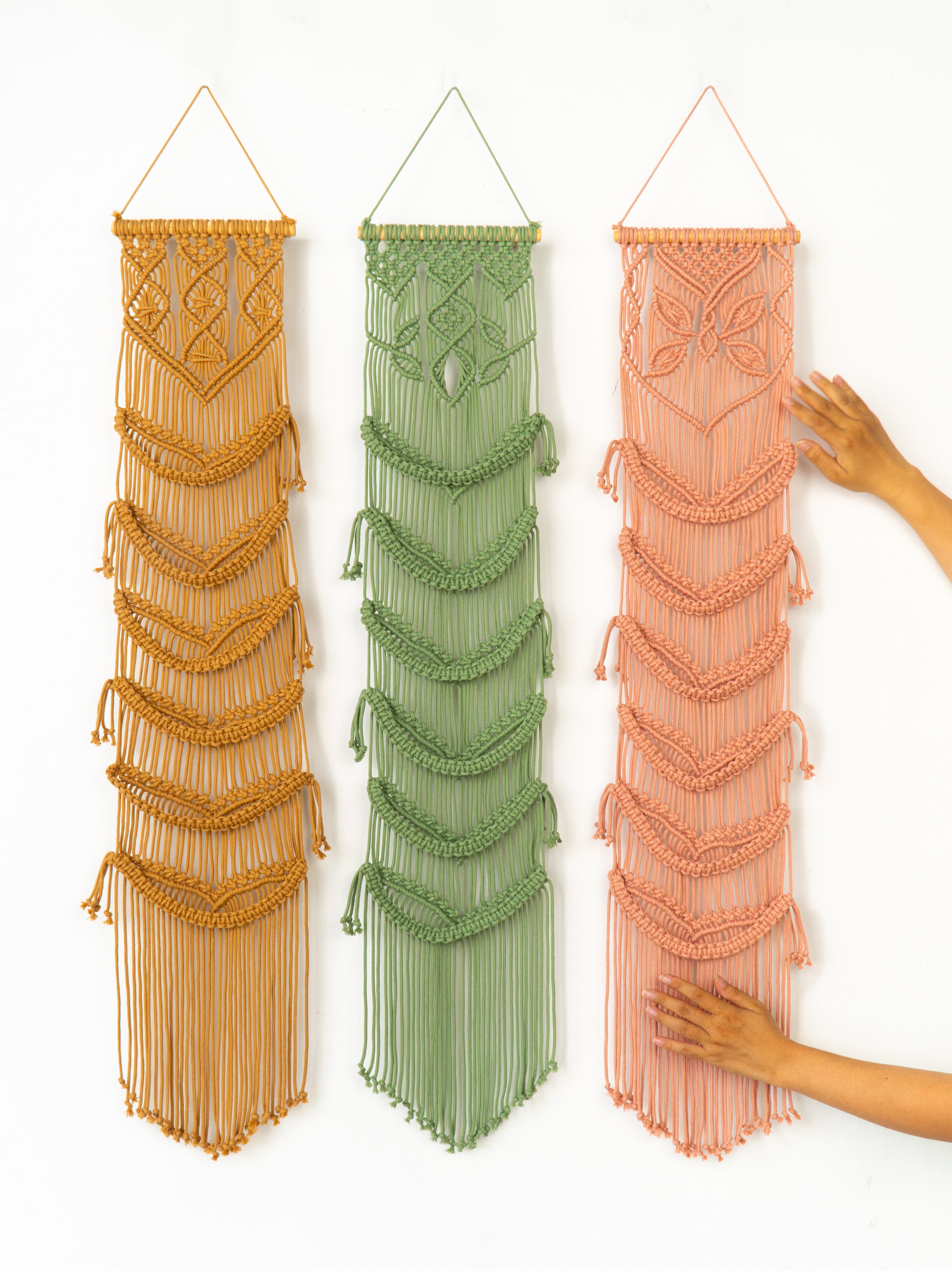 Three macrame wall hangings in gold, green, and peach on a white background.