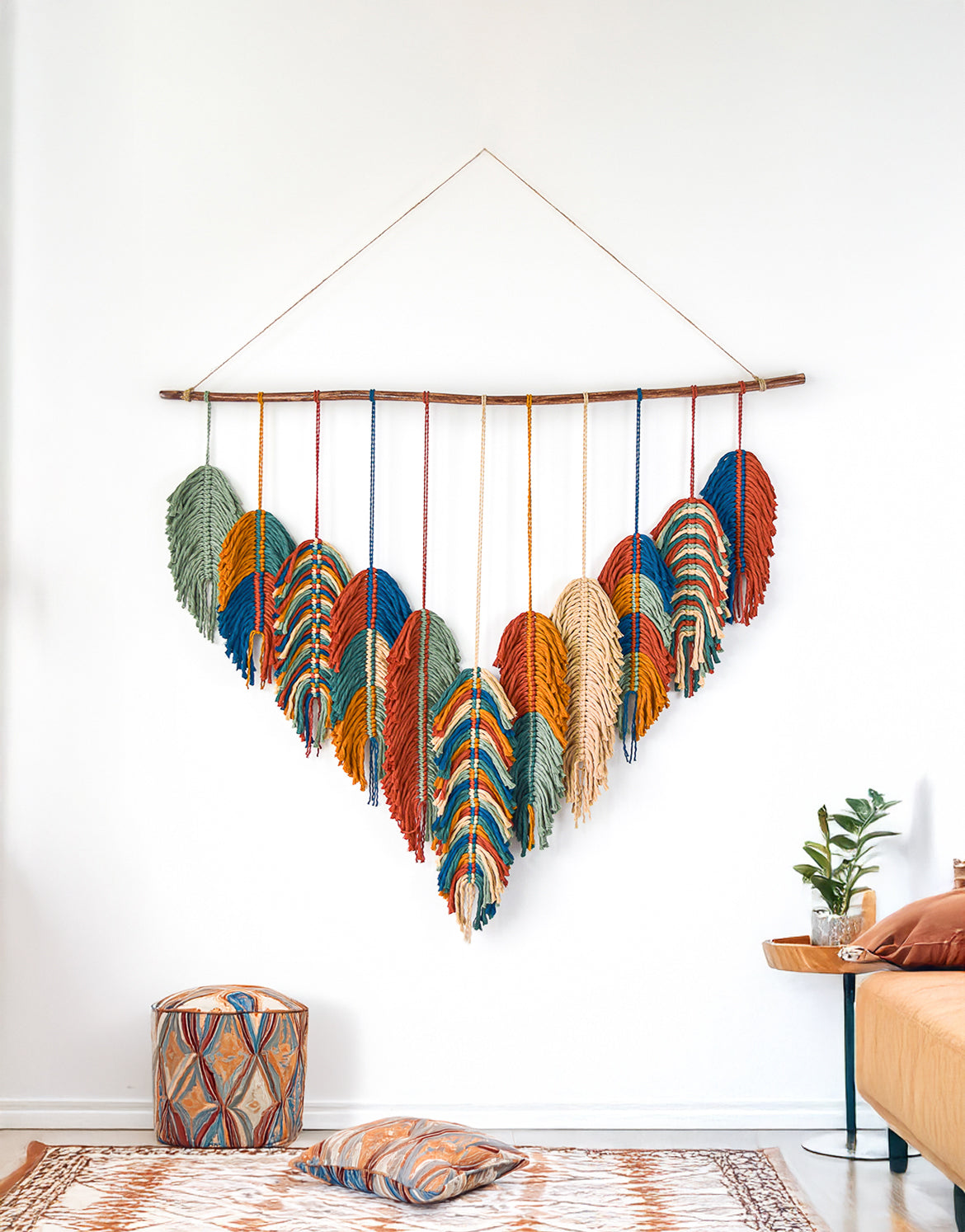 Colorful feather wall hanging in a room with a plant and small table.