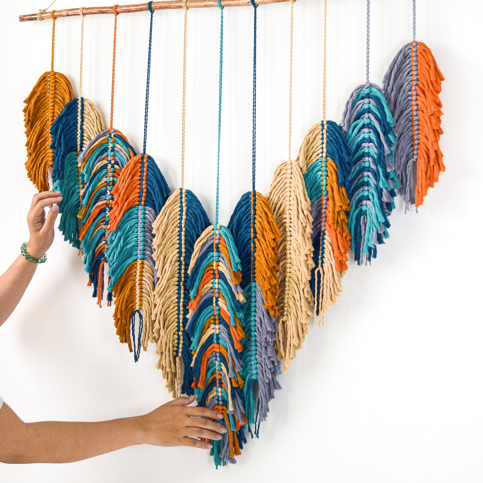 Colorful yarn feather decorations hanging on a wooden rod against a white background.