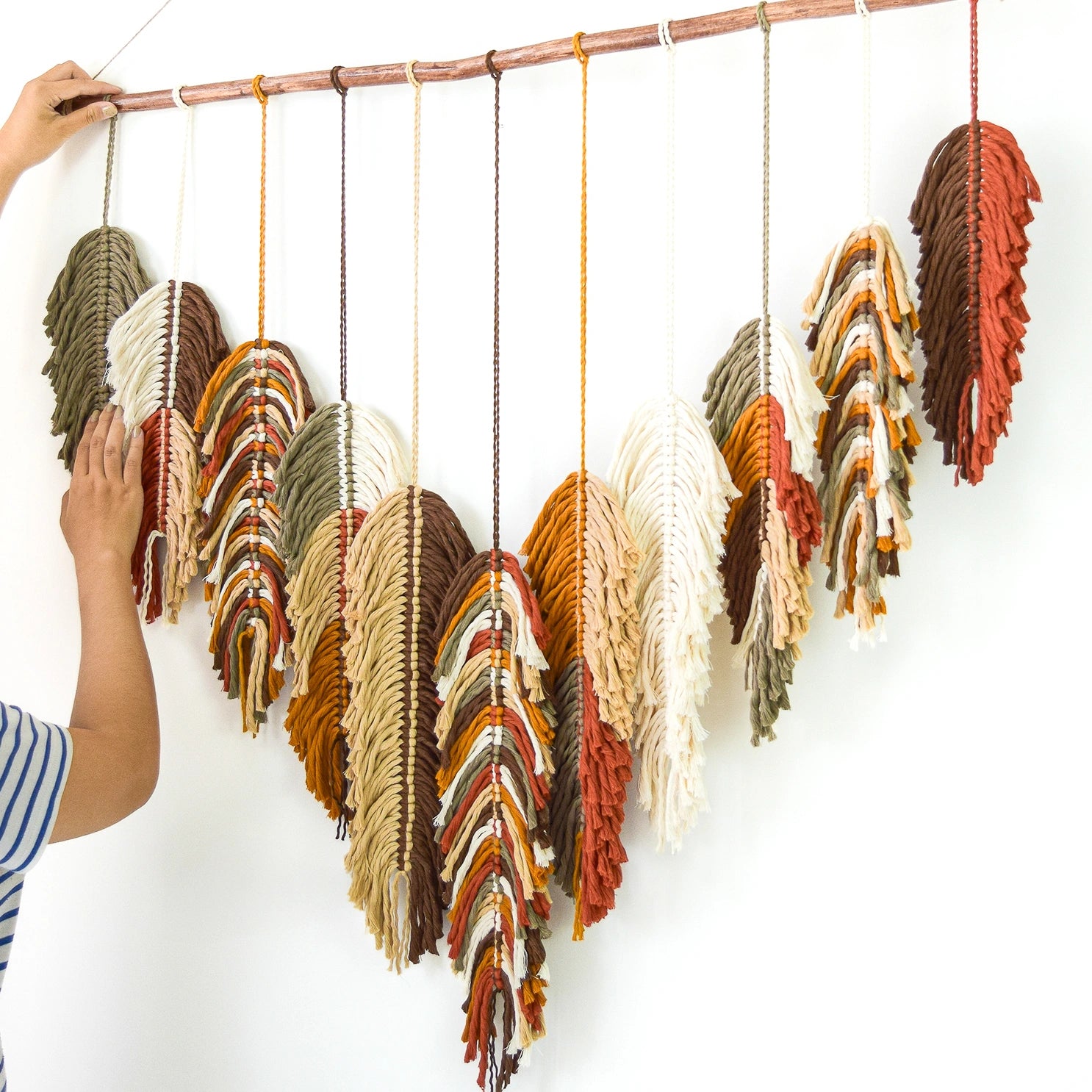 Colorful woven feather decorations hanging from a branch against a white wall.