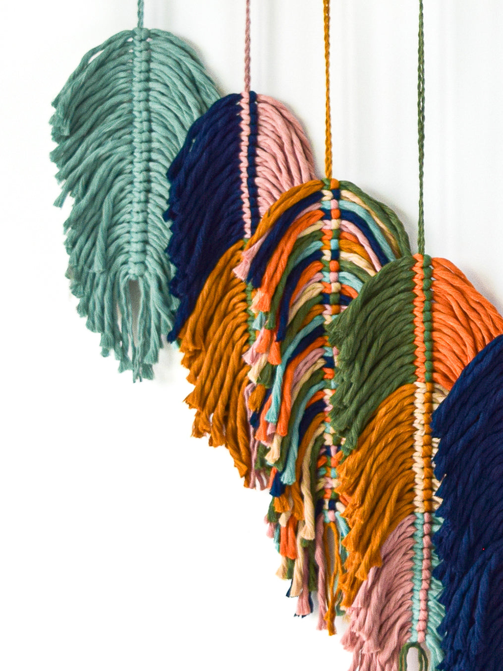 Colorful macrame wall hanging with various shades of green, blue, orange, and pink on a white background.