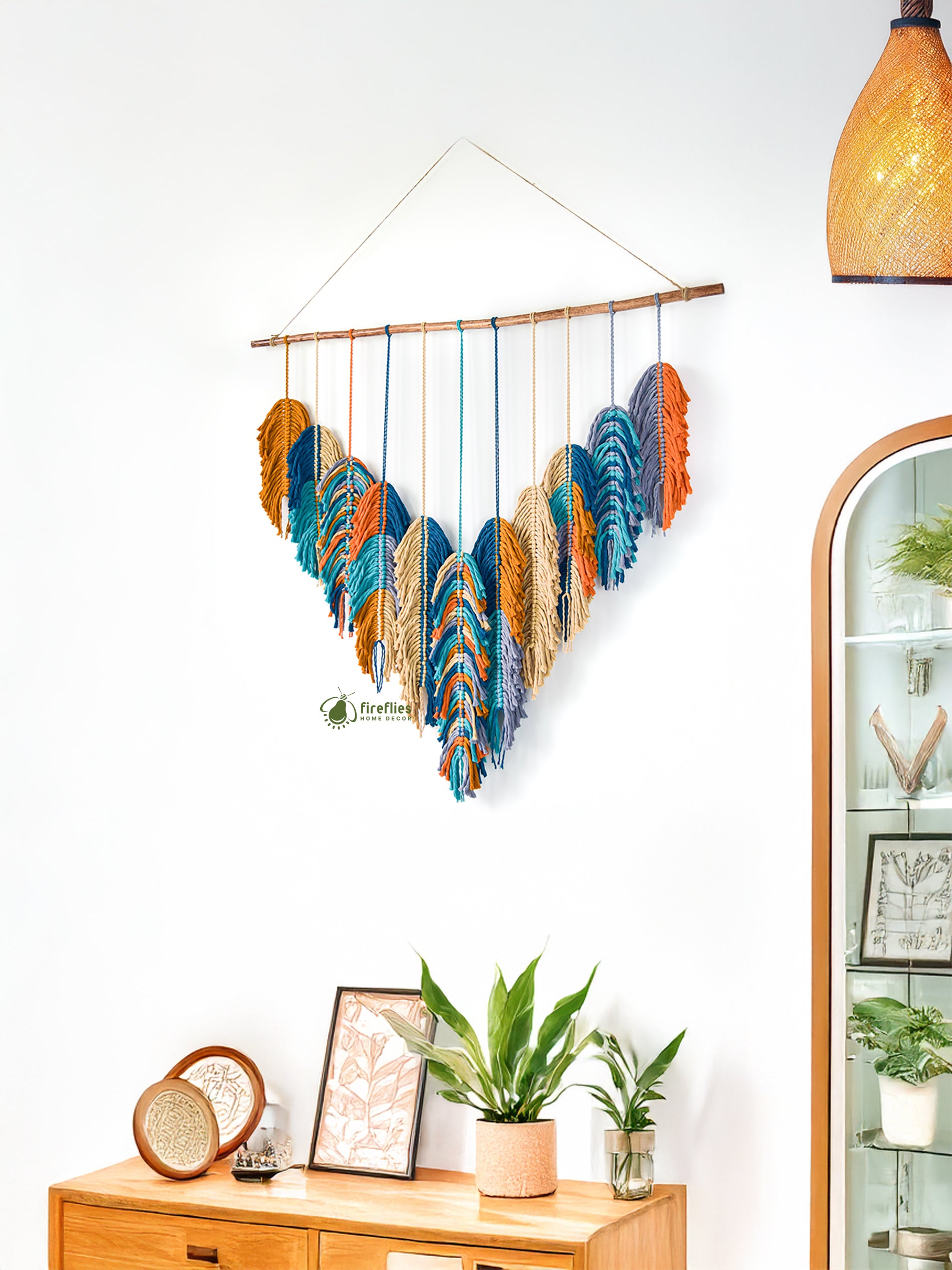 Decorative wall hanging with colorful feathers on a white wall, with a wooden cabinet and plants in the foreground.