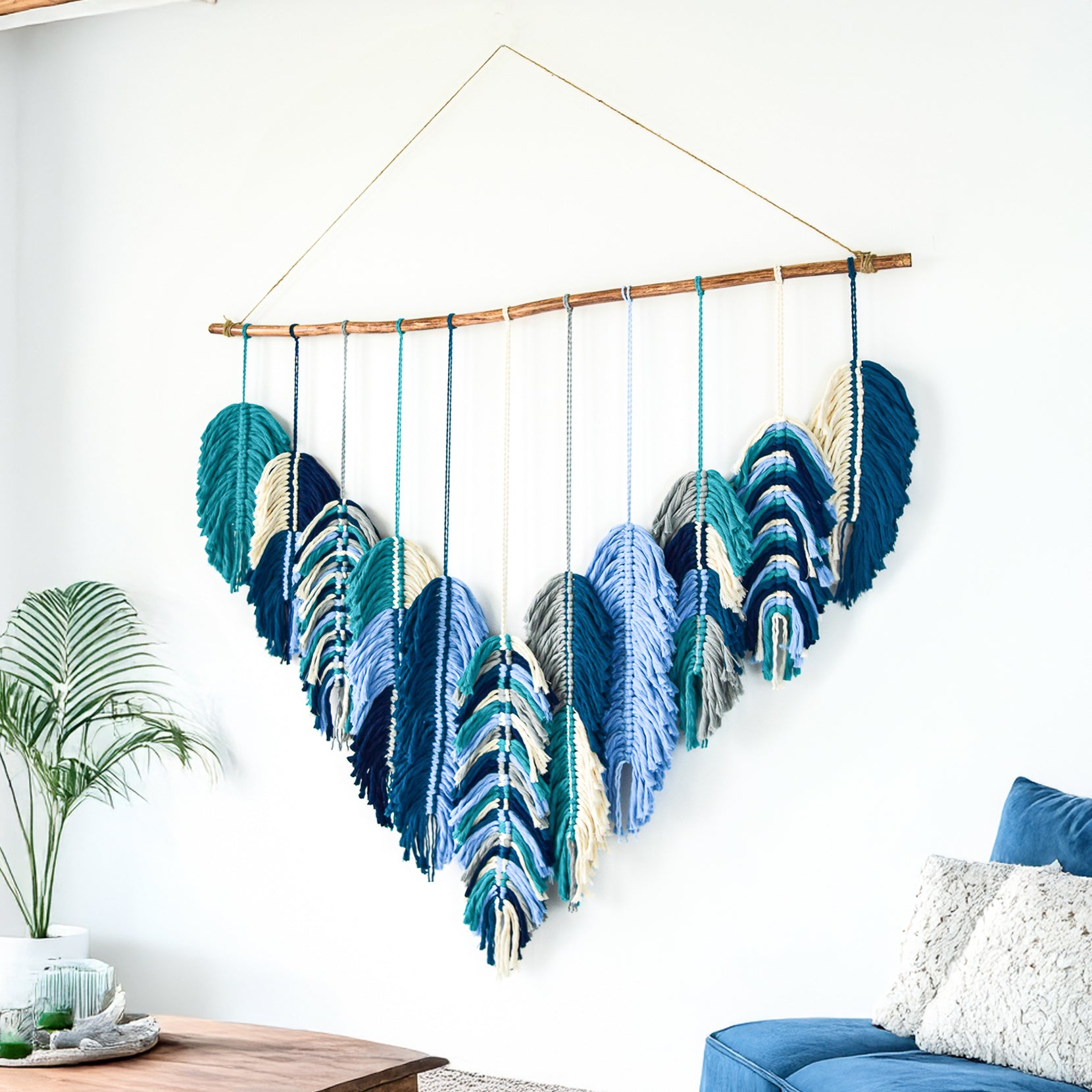 Colorful macrame wall hanging on a wooden rod against a white wall with a lamp and plant in the background.