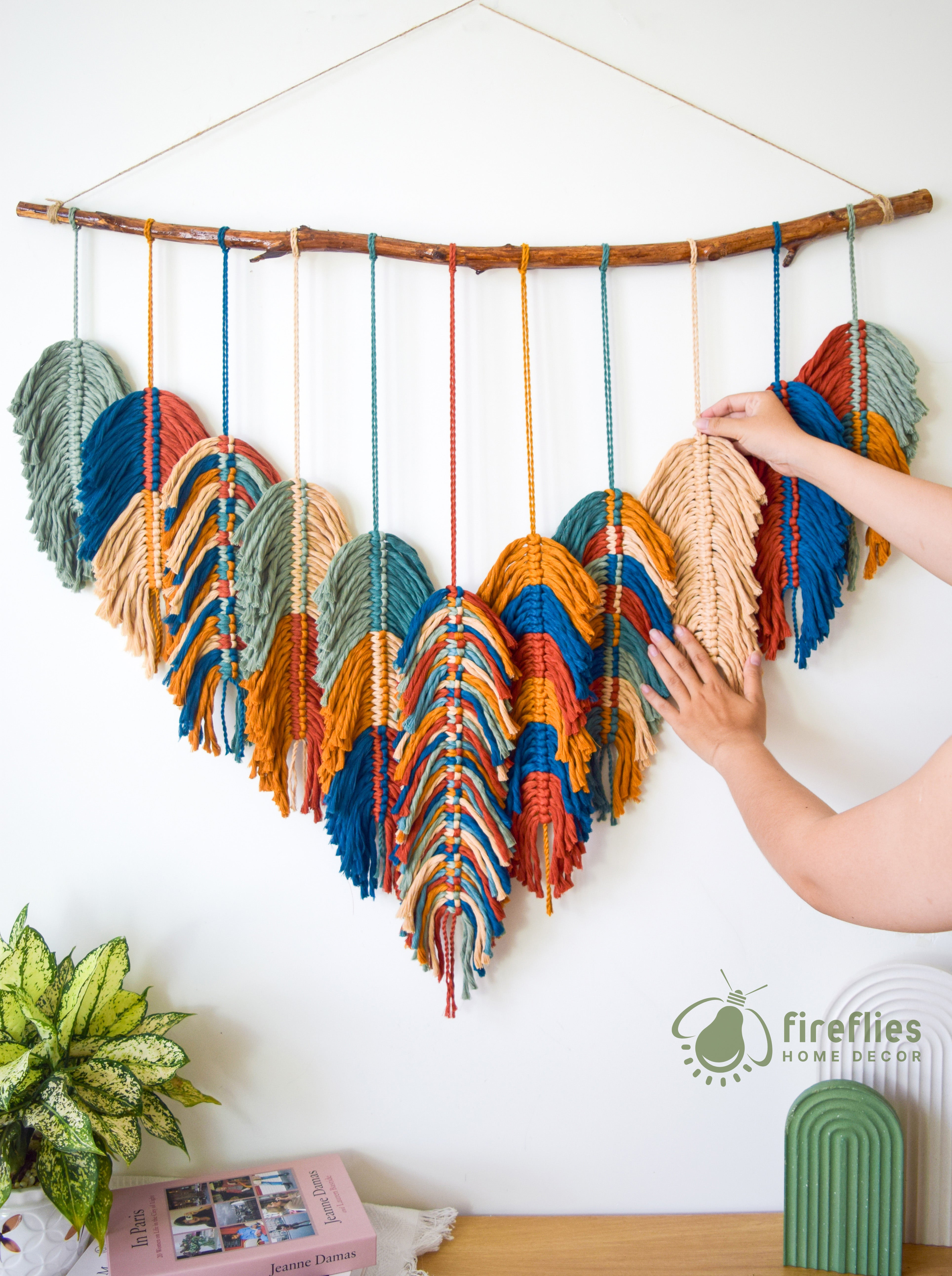 Colorful yarn wall hanging with a branch, hand adjusting it, on a white wall with 'fireflies home decor' branding.