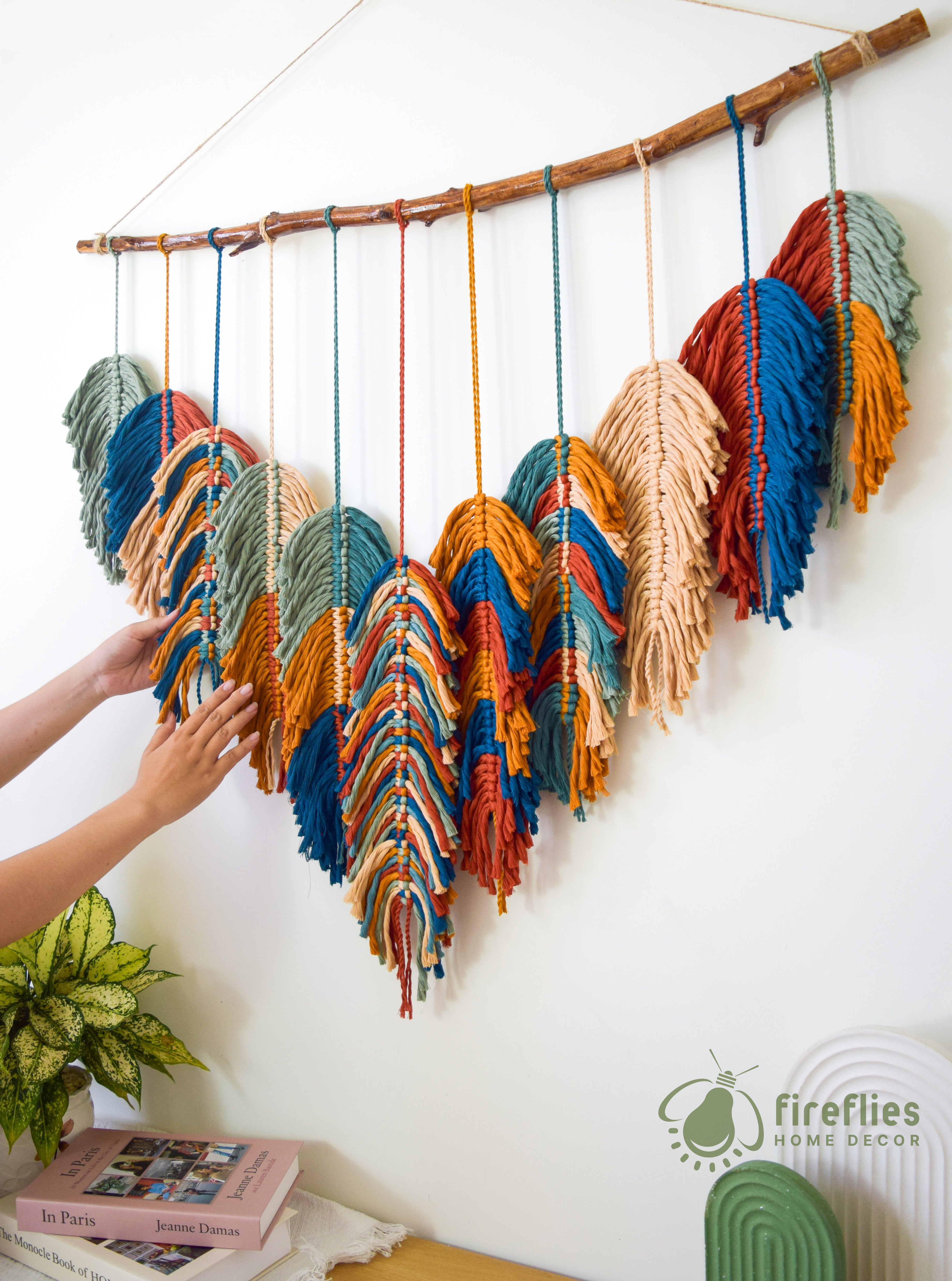 Colorful yarn wall art hanging on a branch with a person's hand touching it, against a white wall.