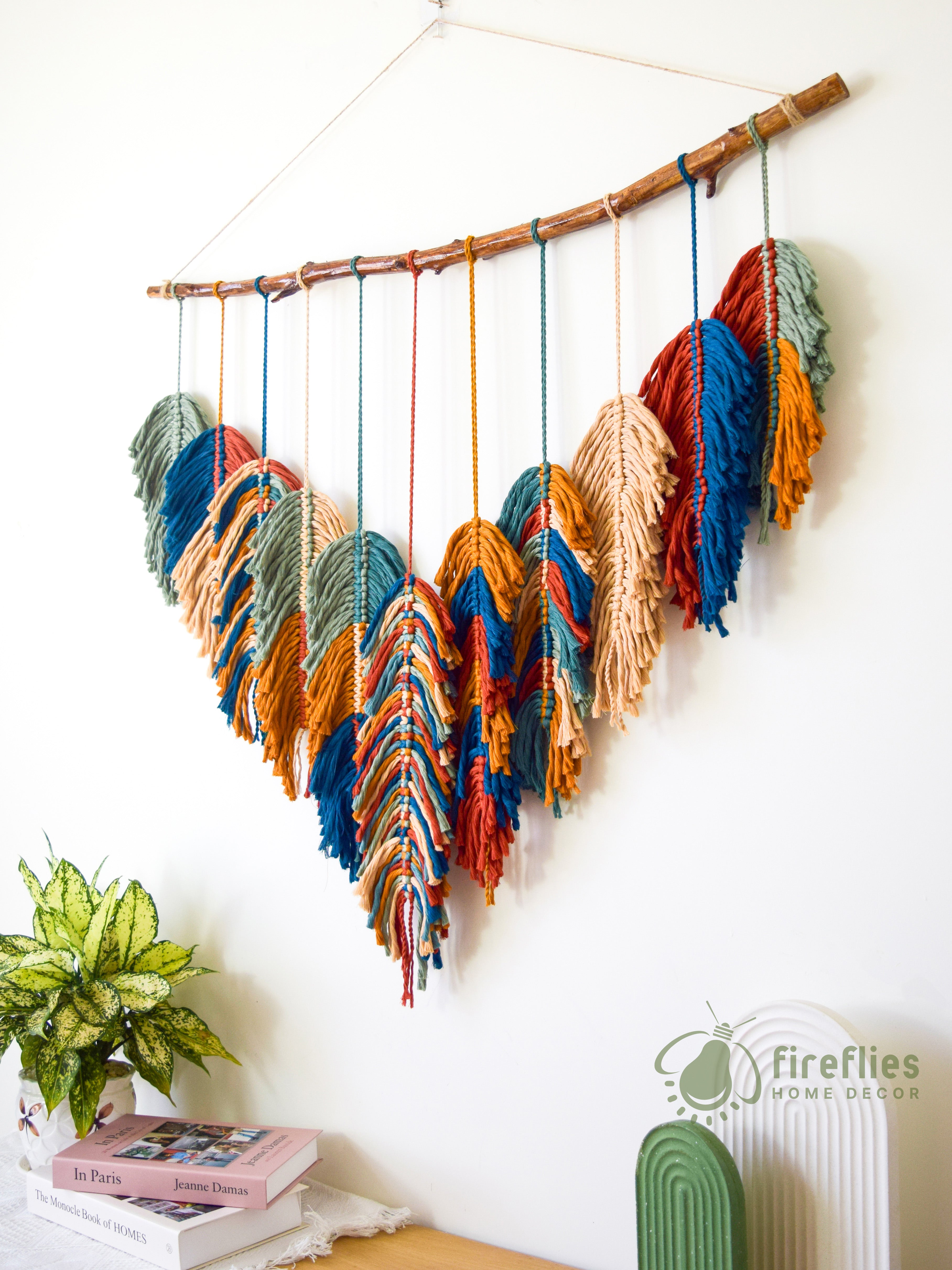 Colorful yarn wall hanging on a branch against a white wall with books and a plant below.